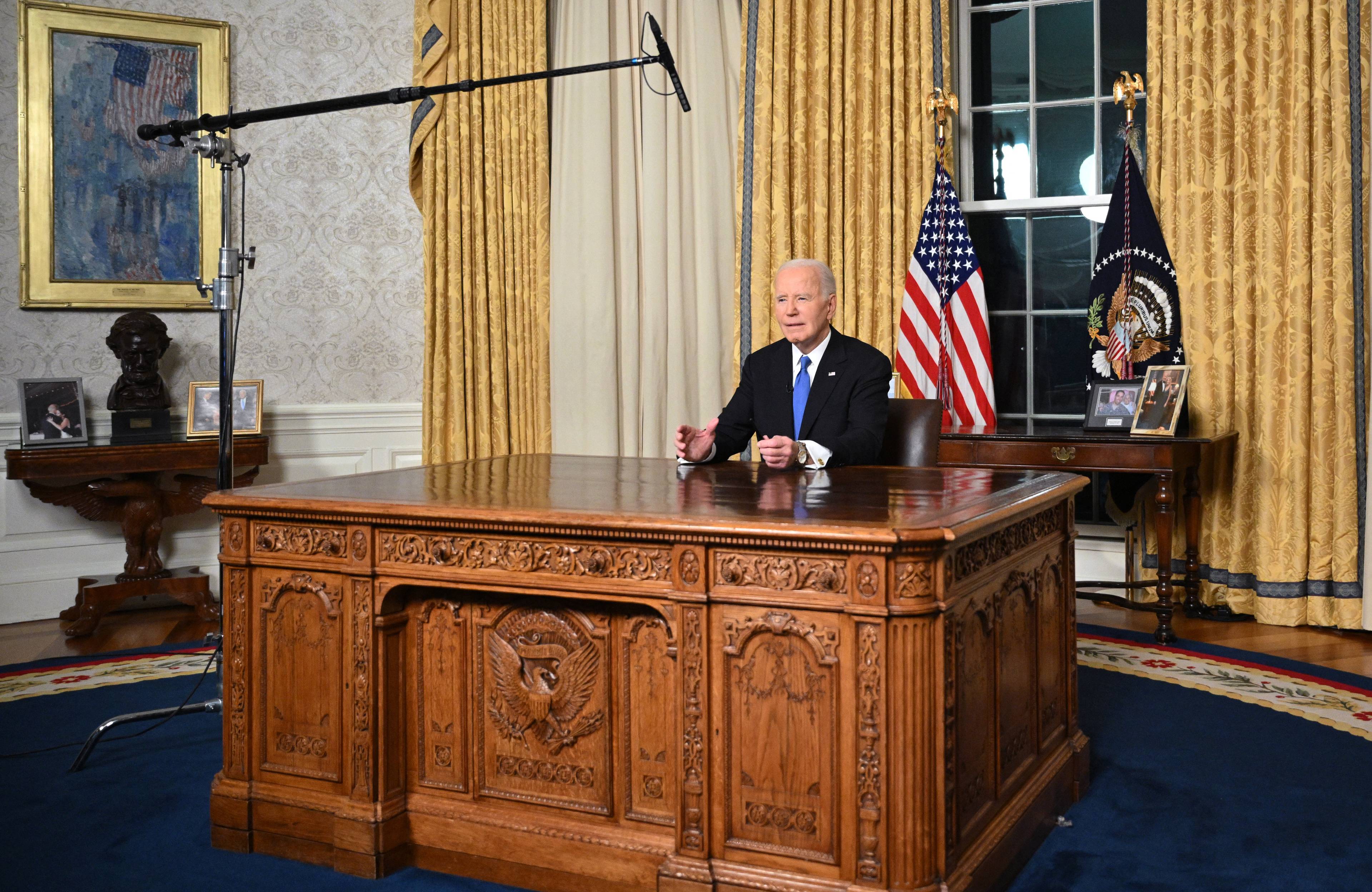Joe Biden siedzi za dużym drewnianym stołem, nad nim mikrofon na wysięgniku, za nim żółte kotary i flaga Stanów Zjednoczonych