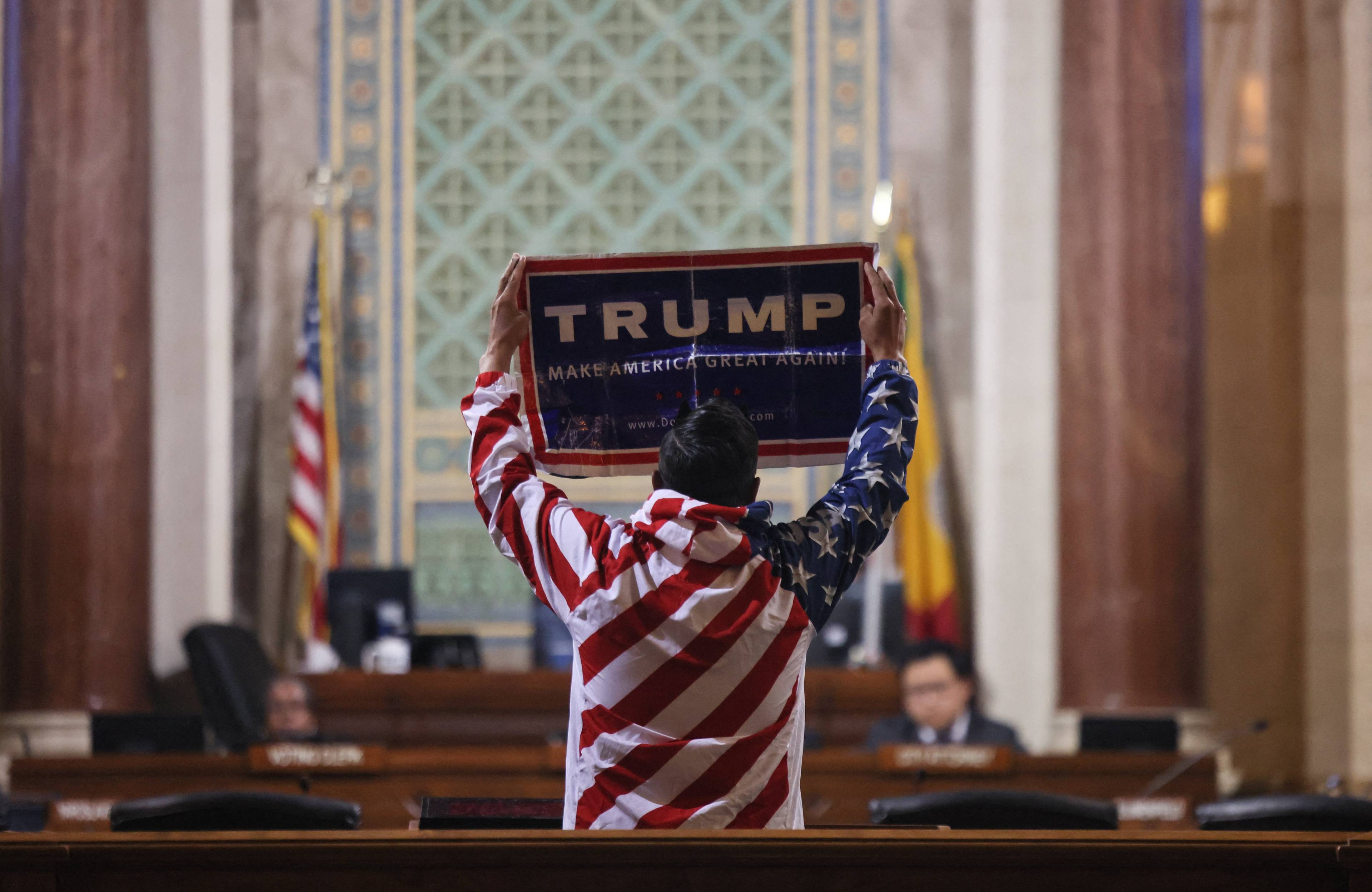 Mężczyzna w koszuli ze wzorem amerykańskiej flagi trzyma nad głową plakat: Trump. Make America Great Again