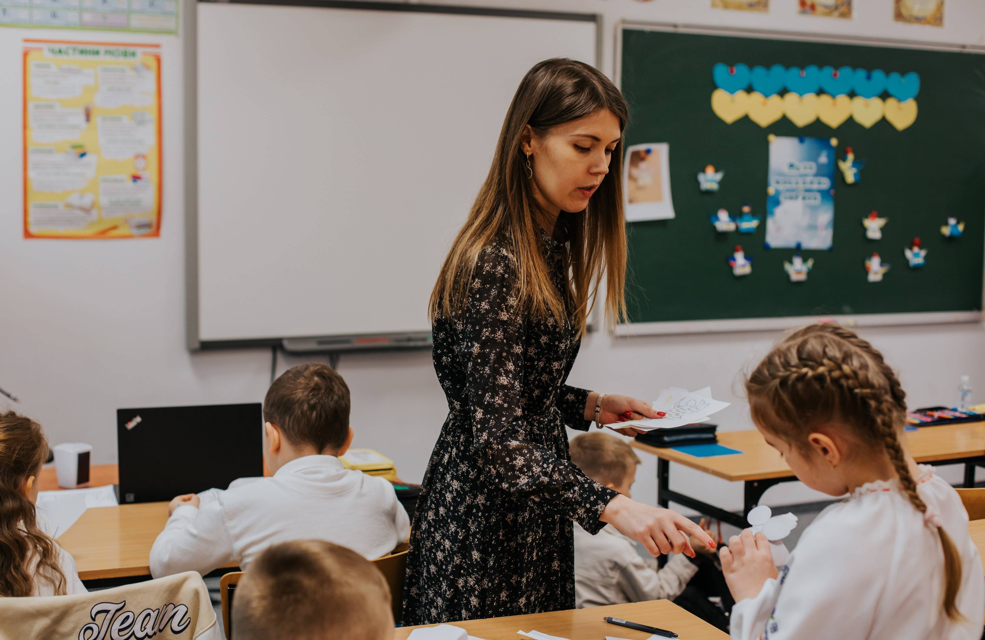 Na zdjęciu widać młodą kobietę z brązowymi włosami oraz troje dzieci w białych koszulach. Dzieci siedzą za ławkami. Z tyłu kobiety jest zielona tablica, na której z serduszek niebieskich i żółtych jest ułożona flaga Ukrainy