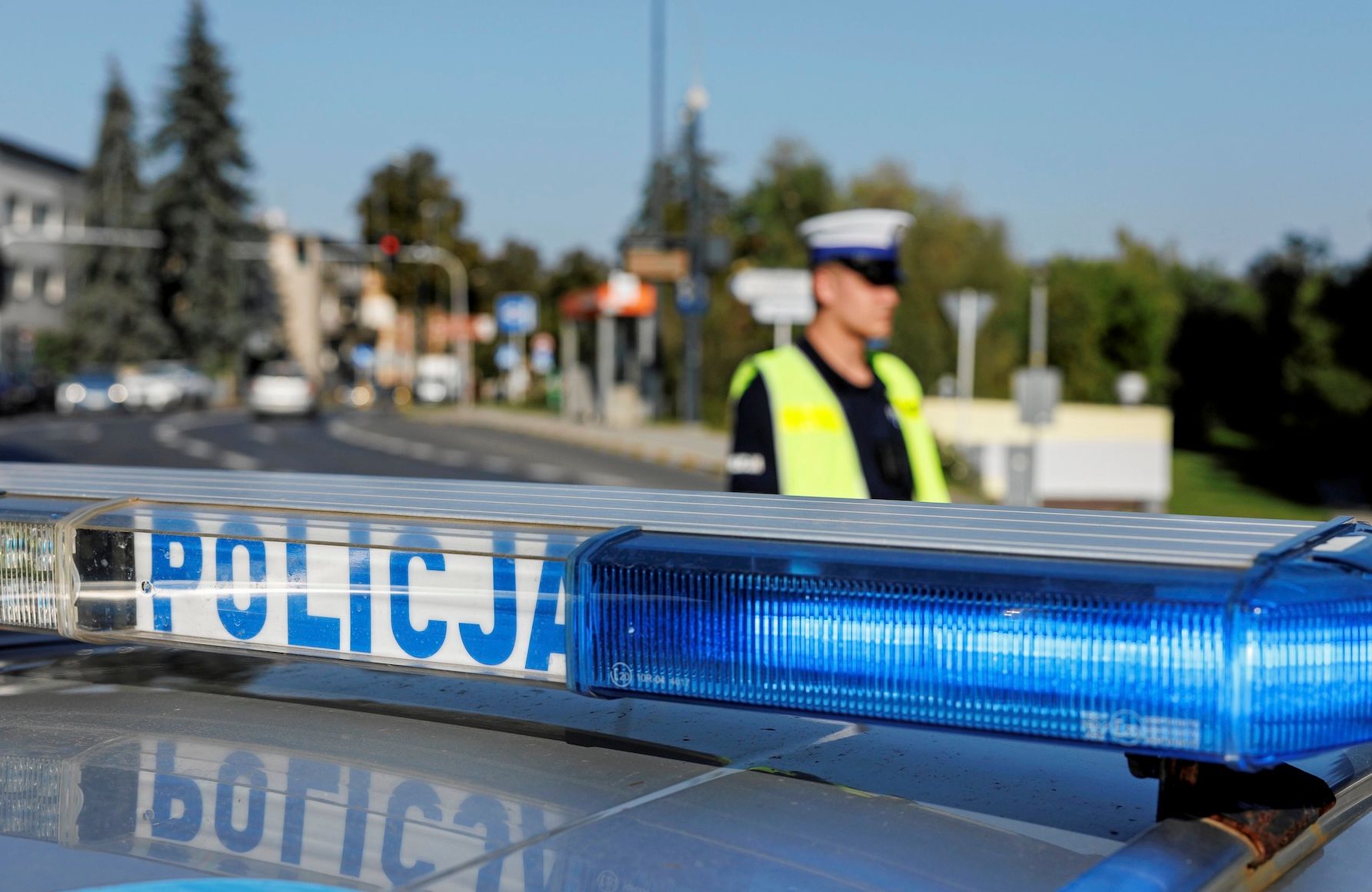 Kogut na dachu policyjnego wozu, w tle policjant w służbowej czapce i odblaskowej kamizelce
