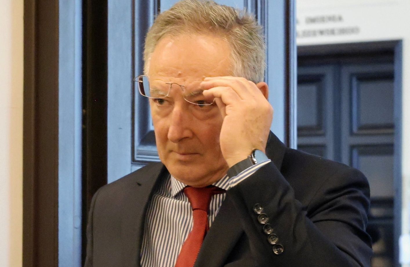 Bartłomiej Sienkiewicz w ciemnym garniturze, koszuli w niebieskie prążki oraz czerwonym krawacie. Podnosi okulary i spogląda spod nich