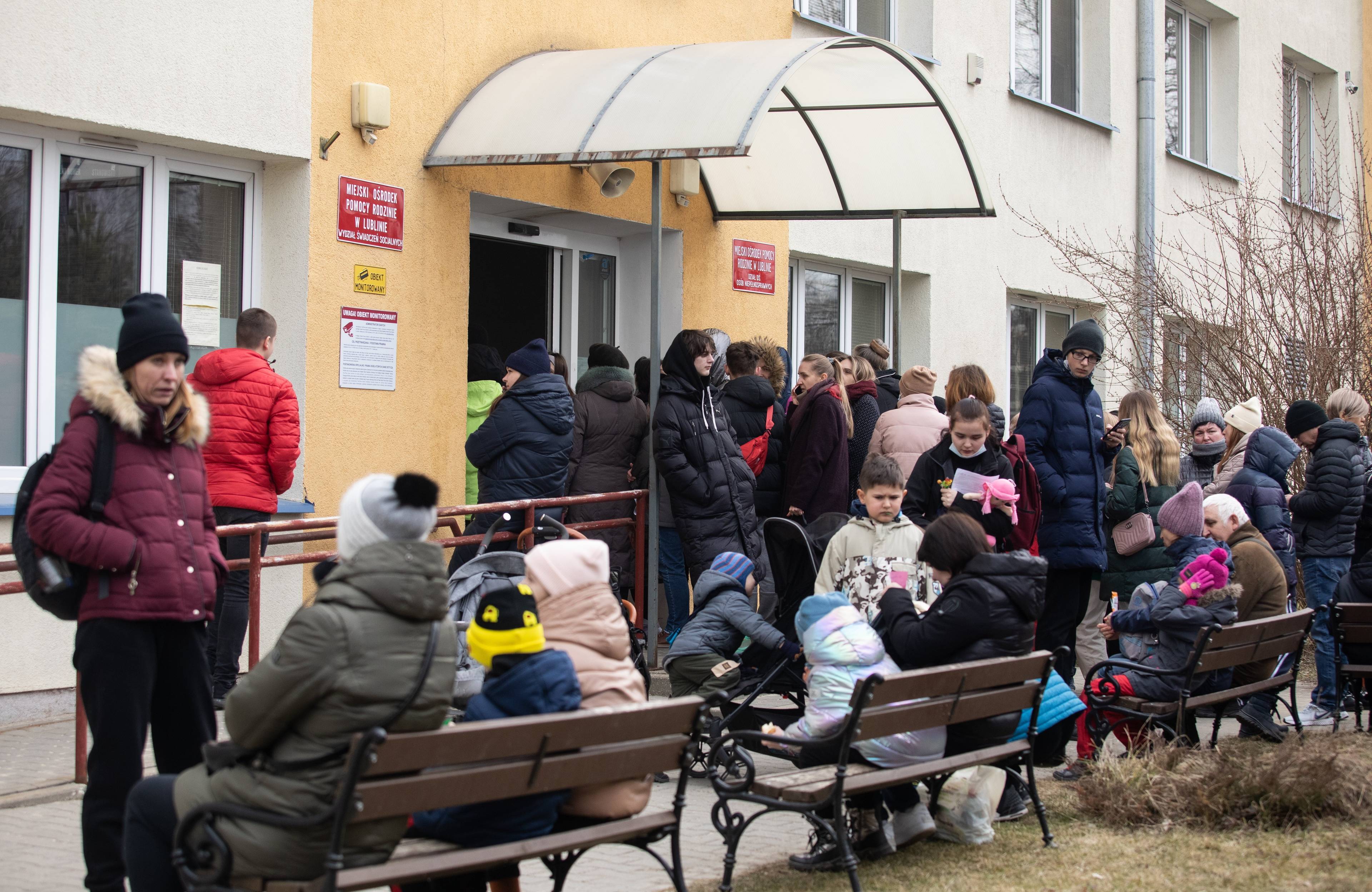 Na zdjęciu widać ludzi, którzy stoją w kolejce obok pomarańczowego budynku, niektórzy siedzą na ławkach obok. Wśród nich są dzieci