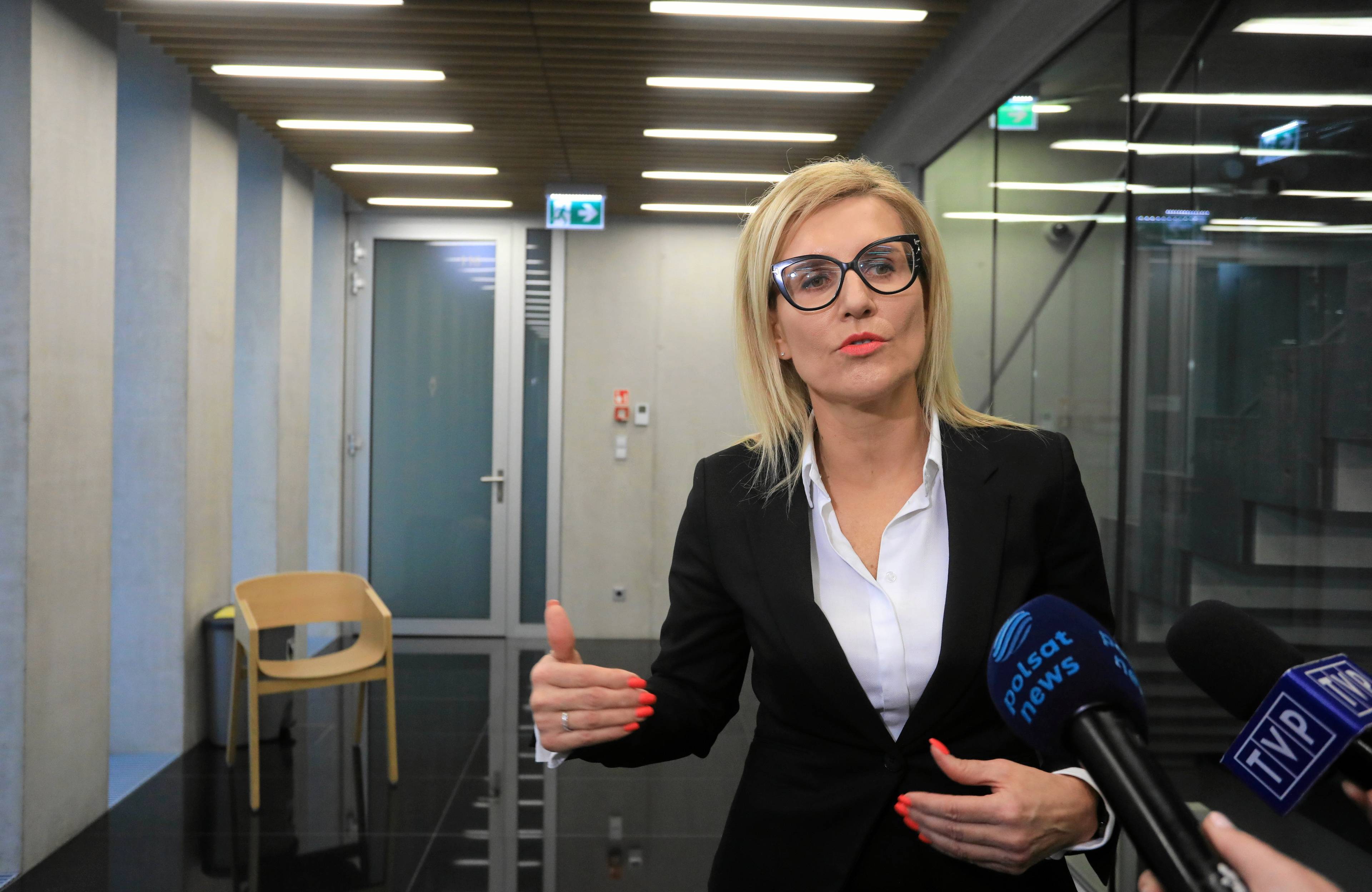 Janowłosa kobieta w jasnej bluzce i ciemnym żakiecie gestykuluje przed mikrofonem