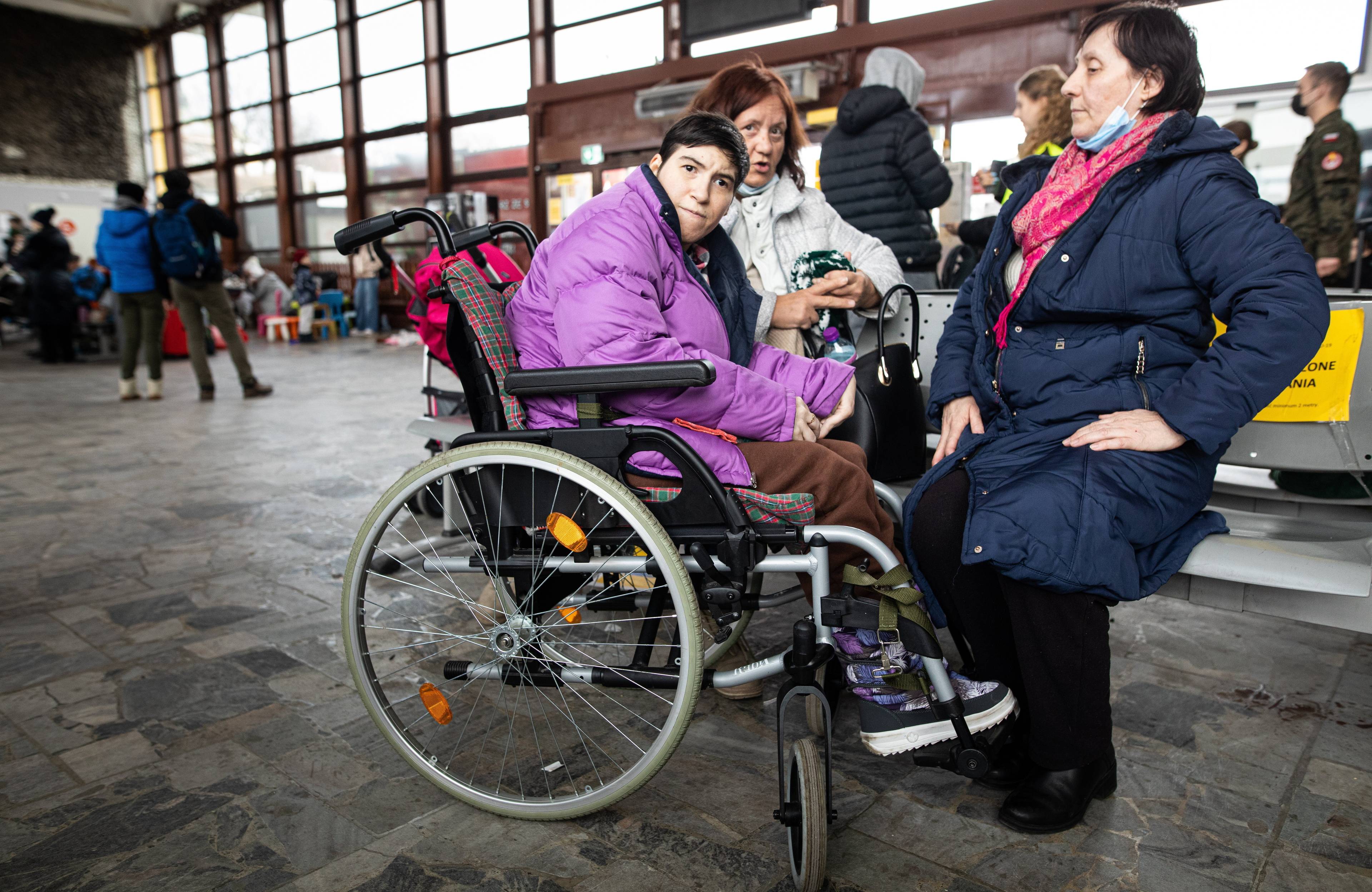 Na zdjęciu jest trzy kobiety. Jedna z nich siedzi w wózku inwalidzkim. Jest ubrana w kurtkę fioletową.
