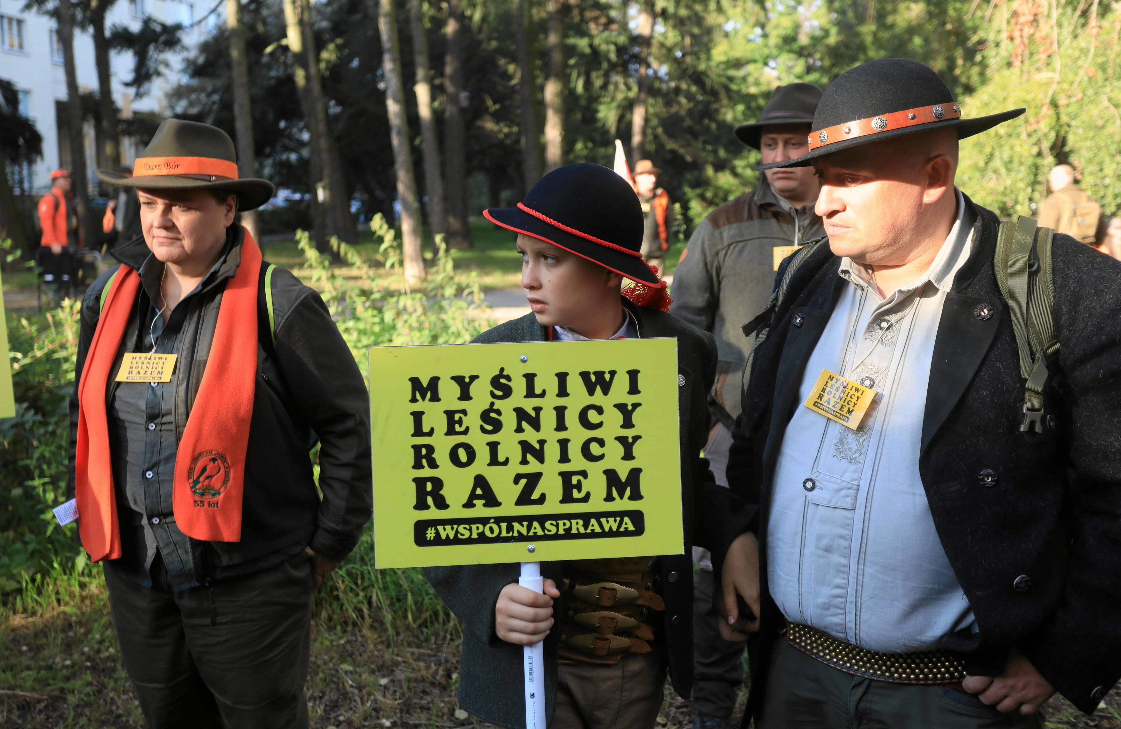 Dwaj męzczyźni i chlopiec w kapeluszach trzumają transparent "Myśliwi, rolnicy razem. Wspólna sprawa"