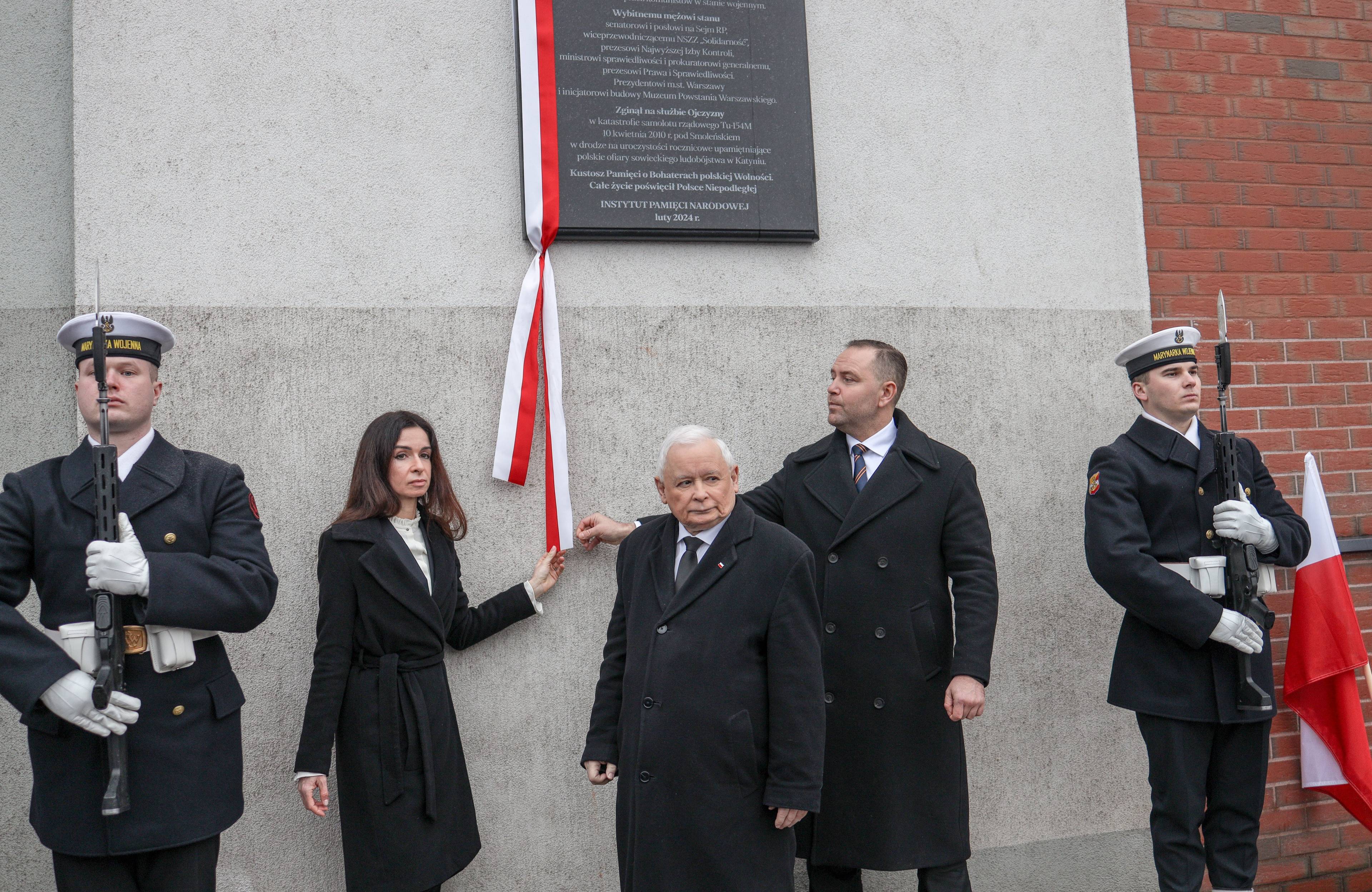 Kilka osób stoi pod wmurowaną w ścianę tablicą upamiętniającą