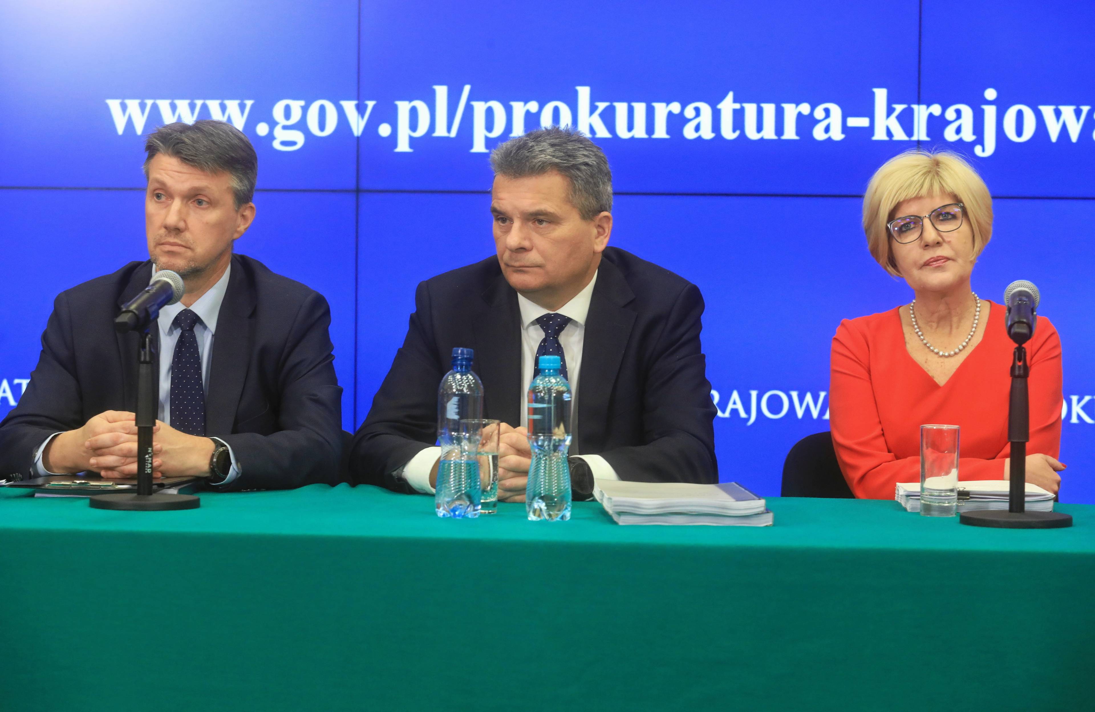 Dwaj mężczyźni i kobieta za stołem pokrytym zielonym suknem. Za nimi logo Prokuratury krajowej