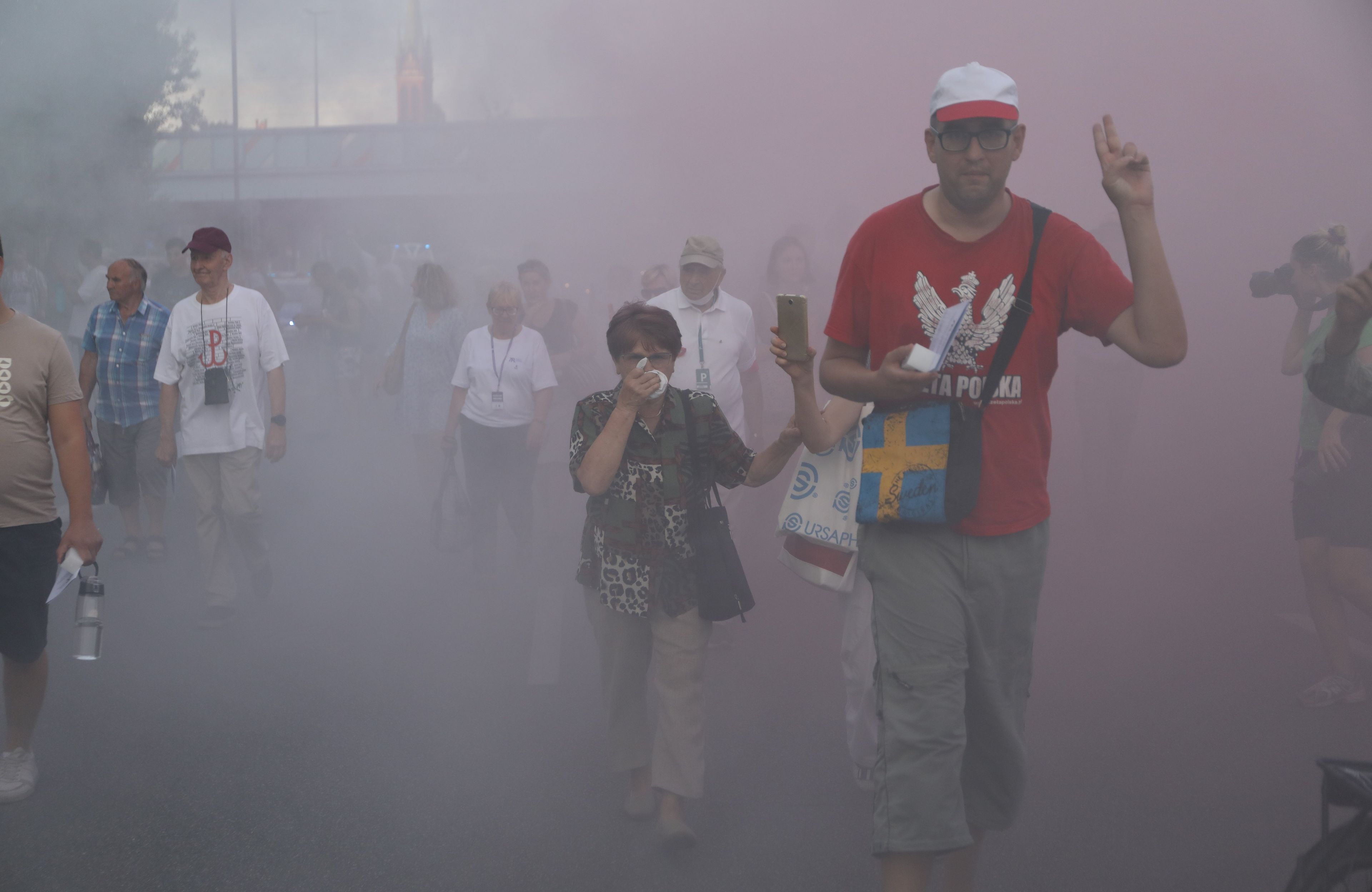z dymu petard wyłania się mężczyzna z orłem na koszulce i kobieta zasłaniająca twarz