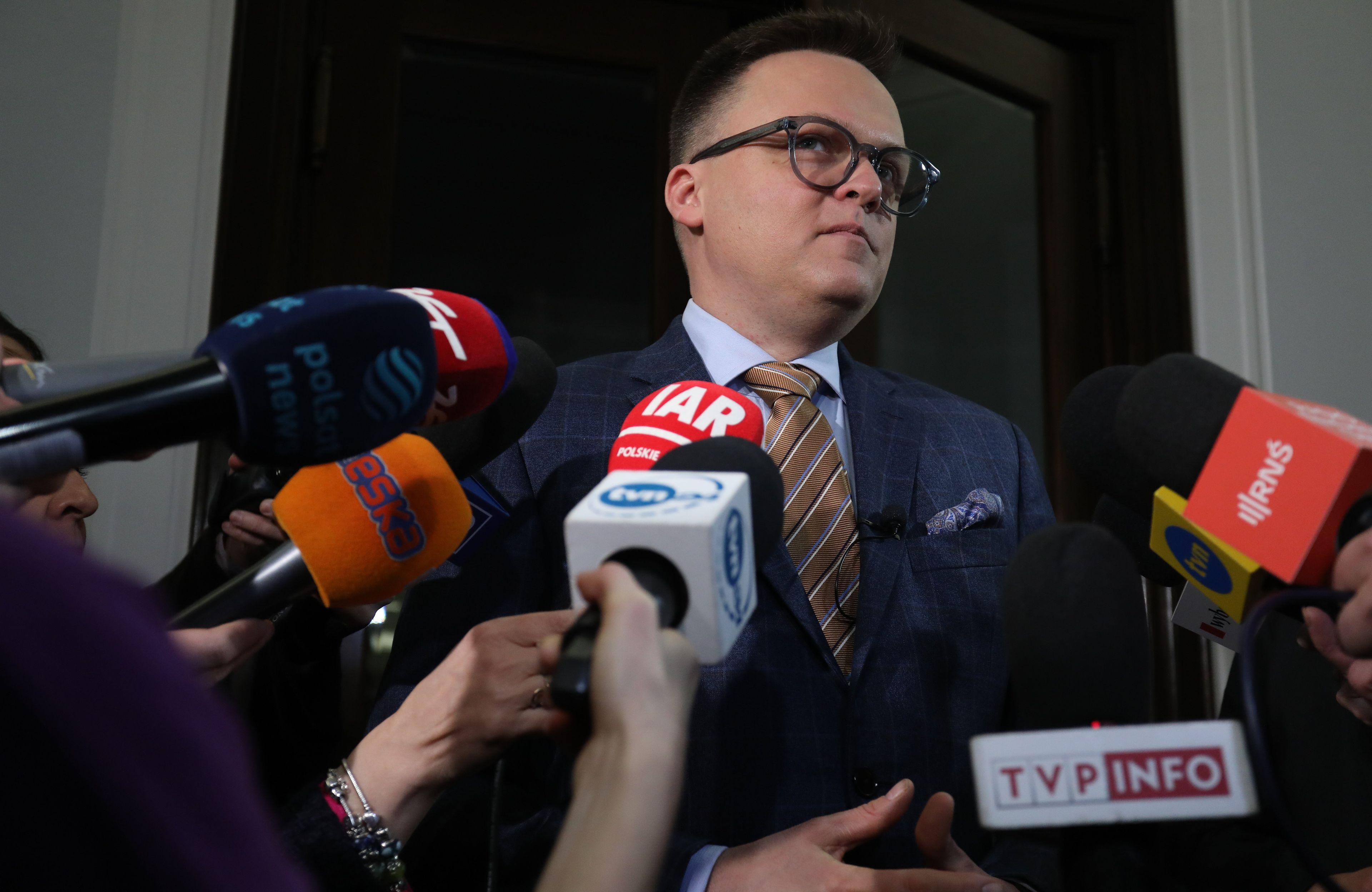 kiść mikrofonów z logami przed 40-latkiem w garniturze i okularach