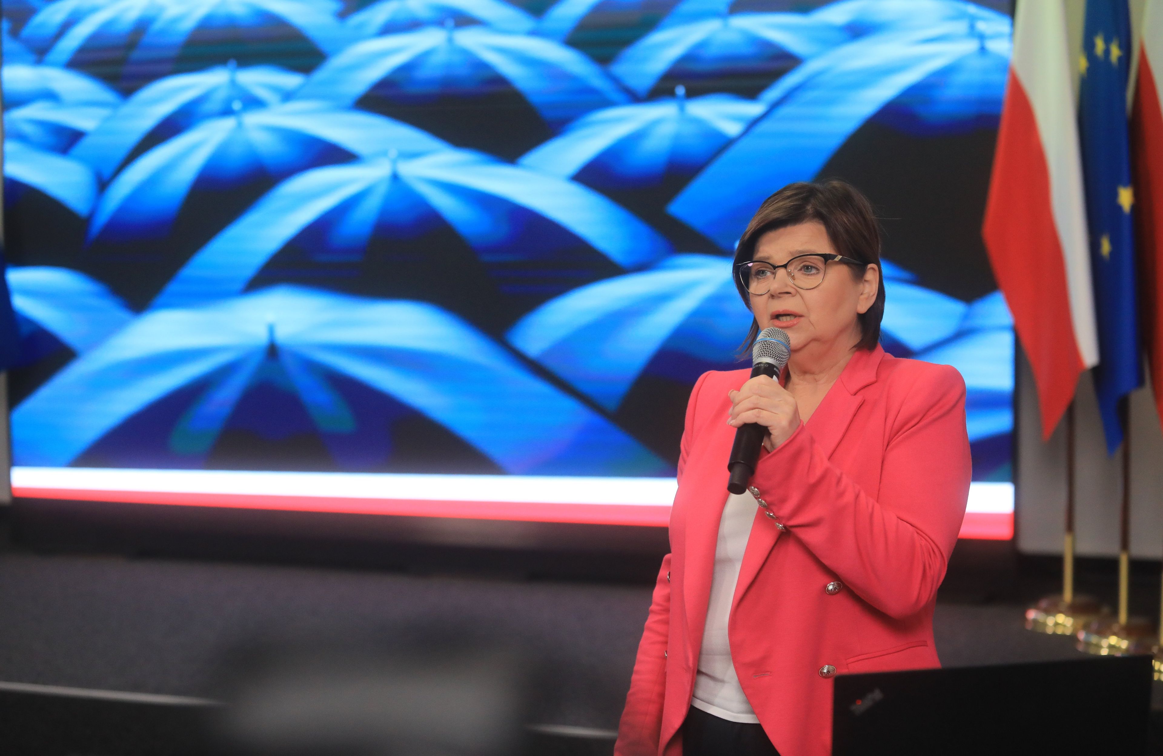 Kobieta w czerwonej marynarce (ministra zdrowia) na tle zdjęcia czarnych parasolek - symbolu protestu kobiet
