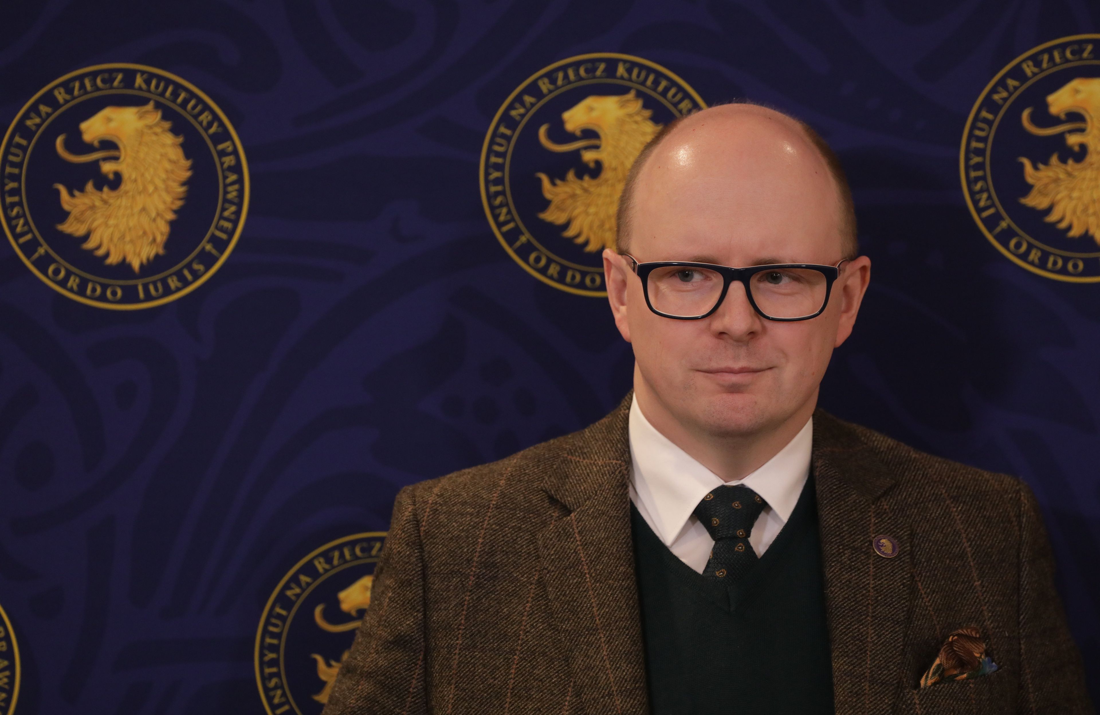 Łysy mężczyzna w okularach na tle logo z napisem Ordo Iuris