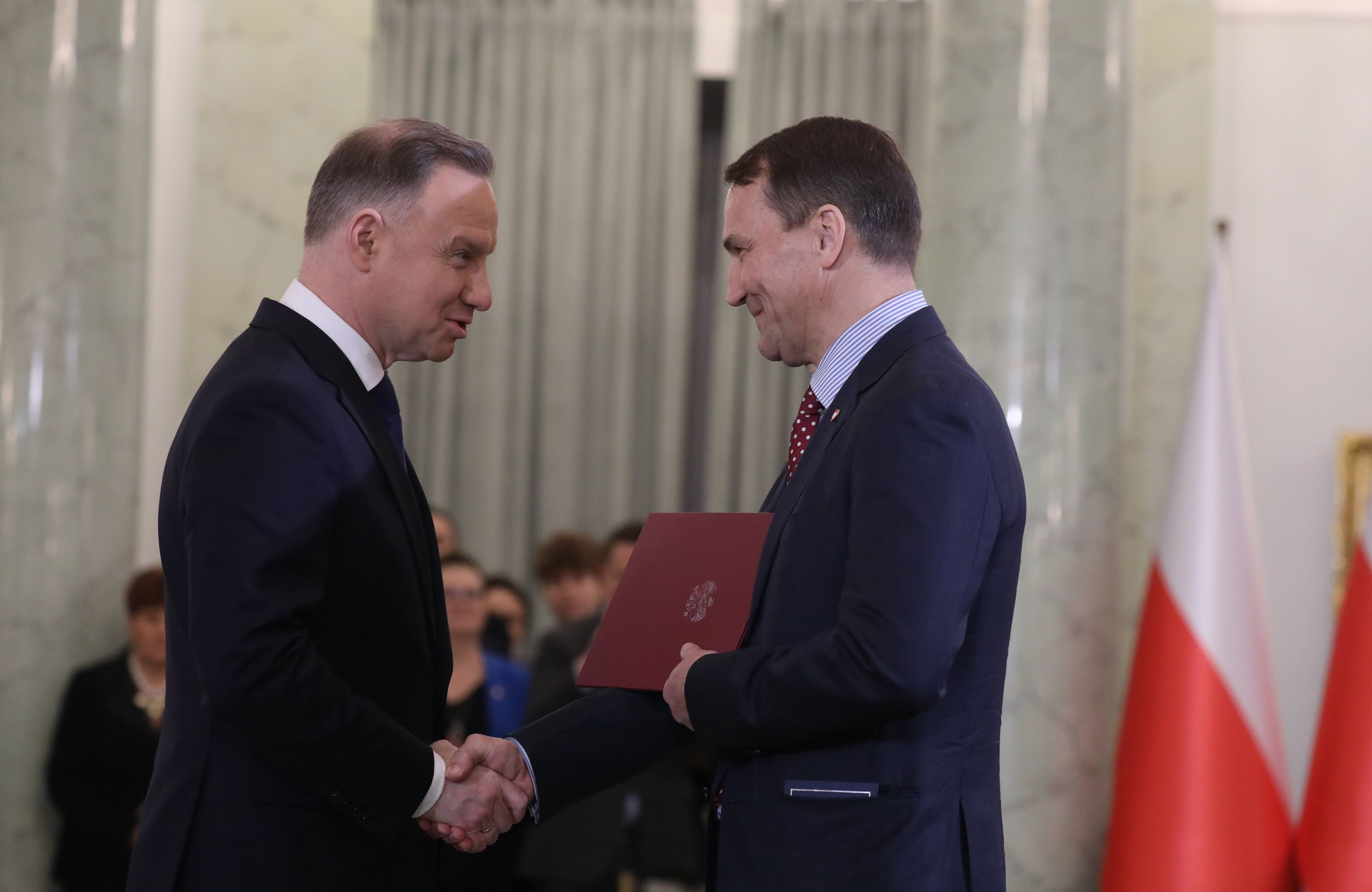 Dwóch panów w garniturach podaje sobie ręce na tle flagi Polski
