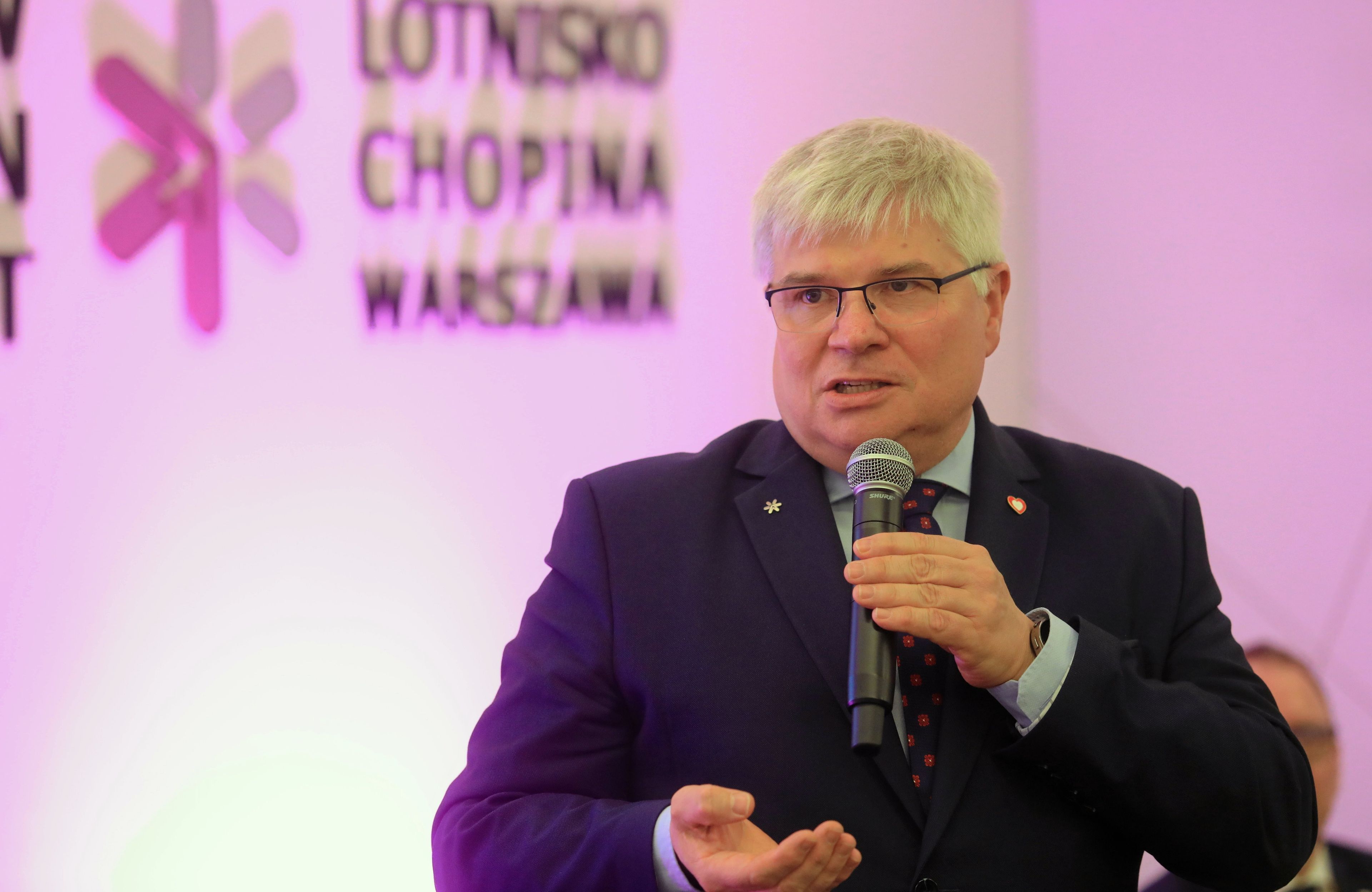 siwy mężczyzna w okularach przemawia z mikrofonem, w tle logo lotniska Chopina