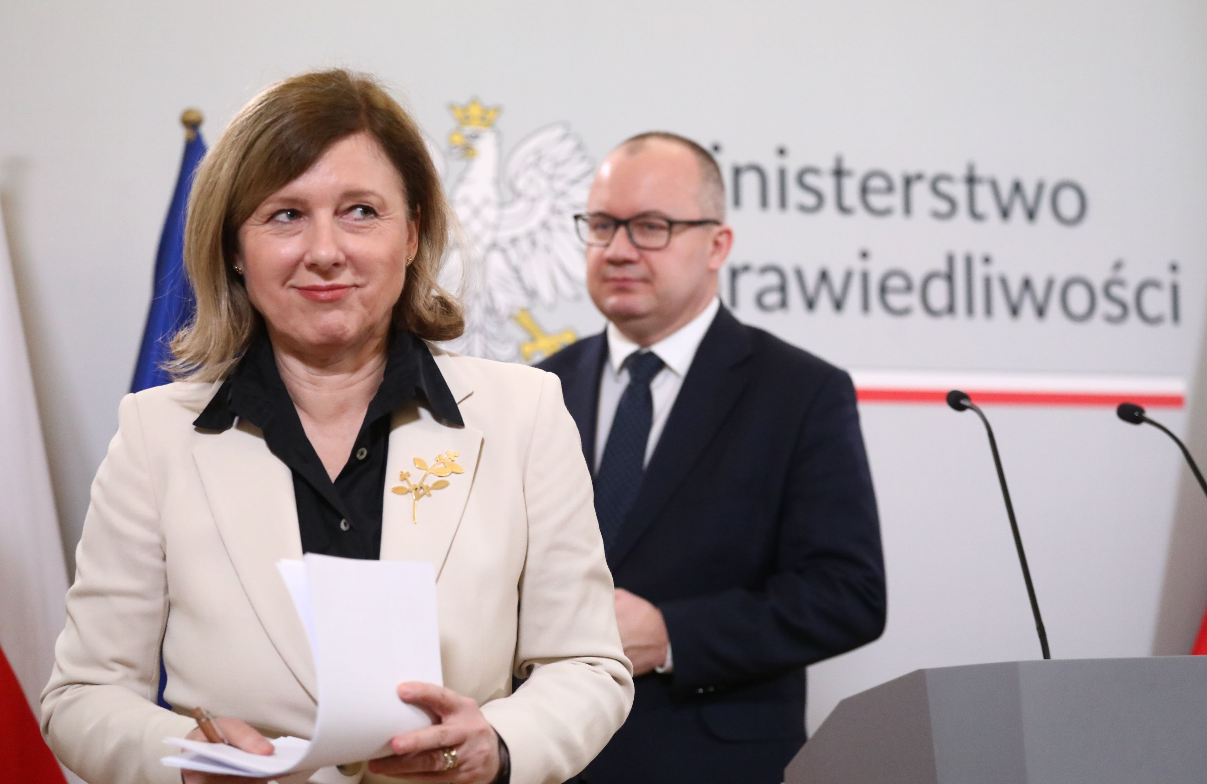 Kobieta w jasnym żakiecie, za nią mężczyzna w garniturze