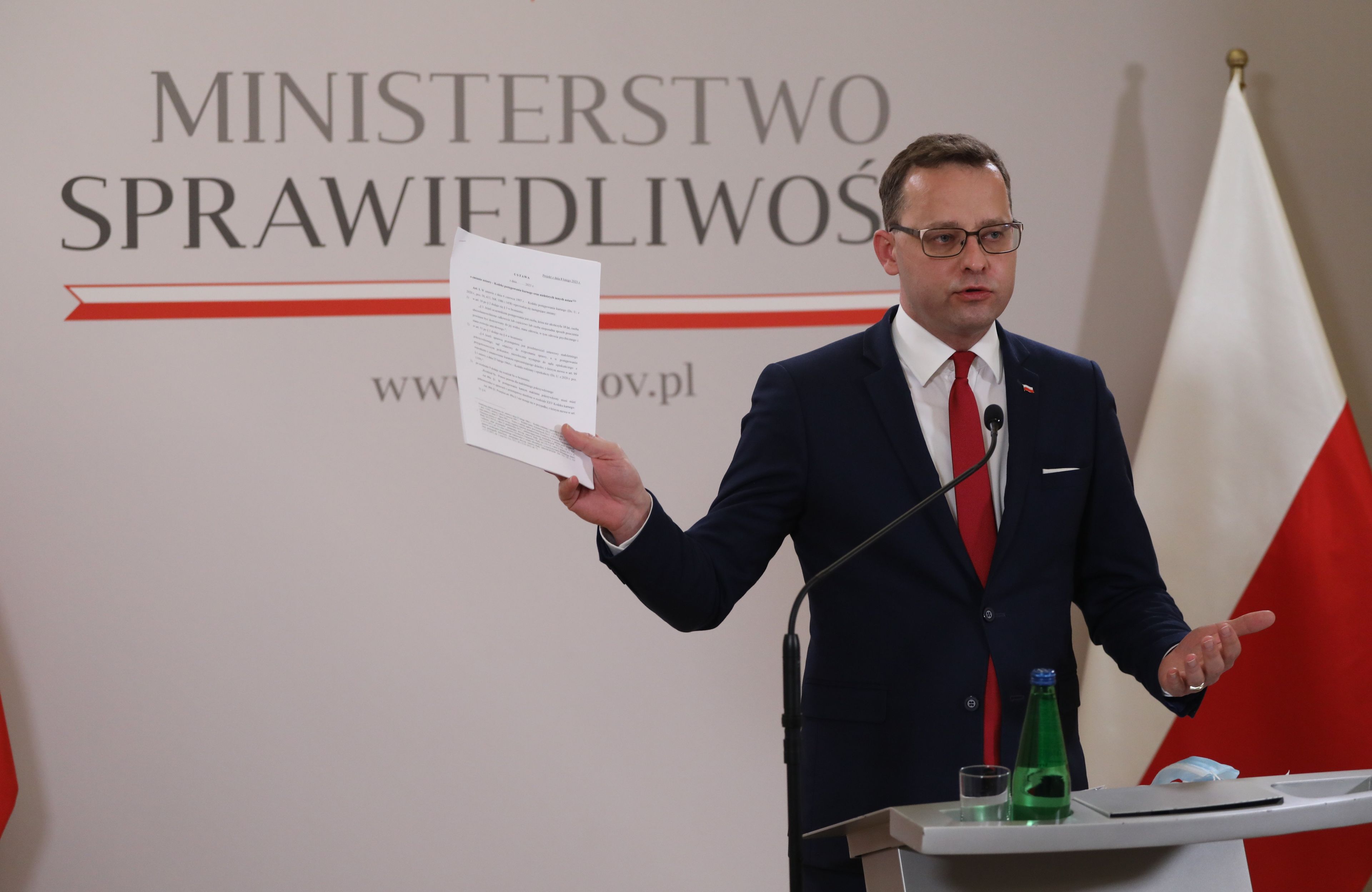Mężczyzna za mównicą trzyma w lewej ręce plik kartek. W tle napis "Ministerstwo Sprawiedliwości"