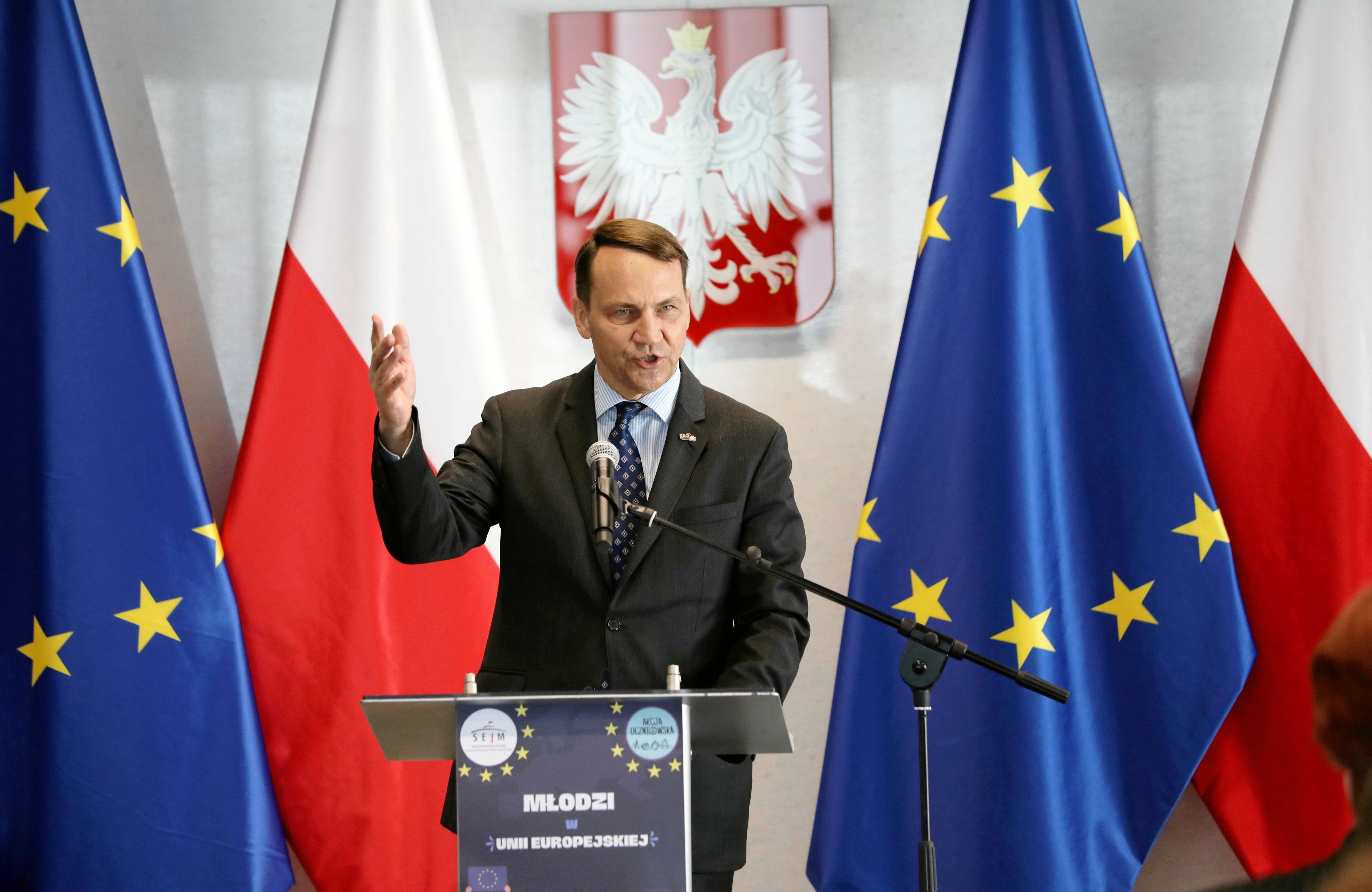 Polityk w garniturze z podniesioną ręką przemawia na tle flag Polski i Unii