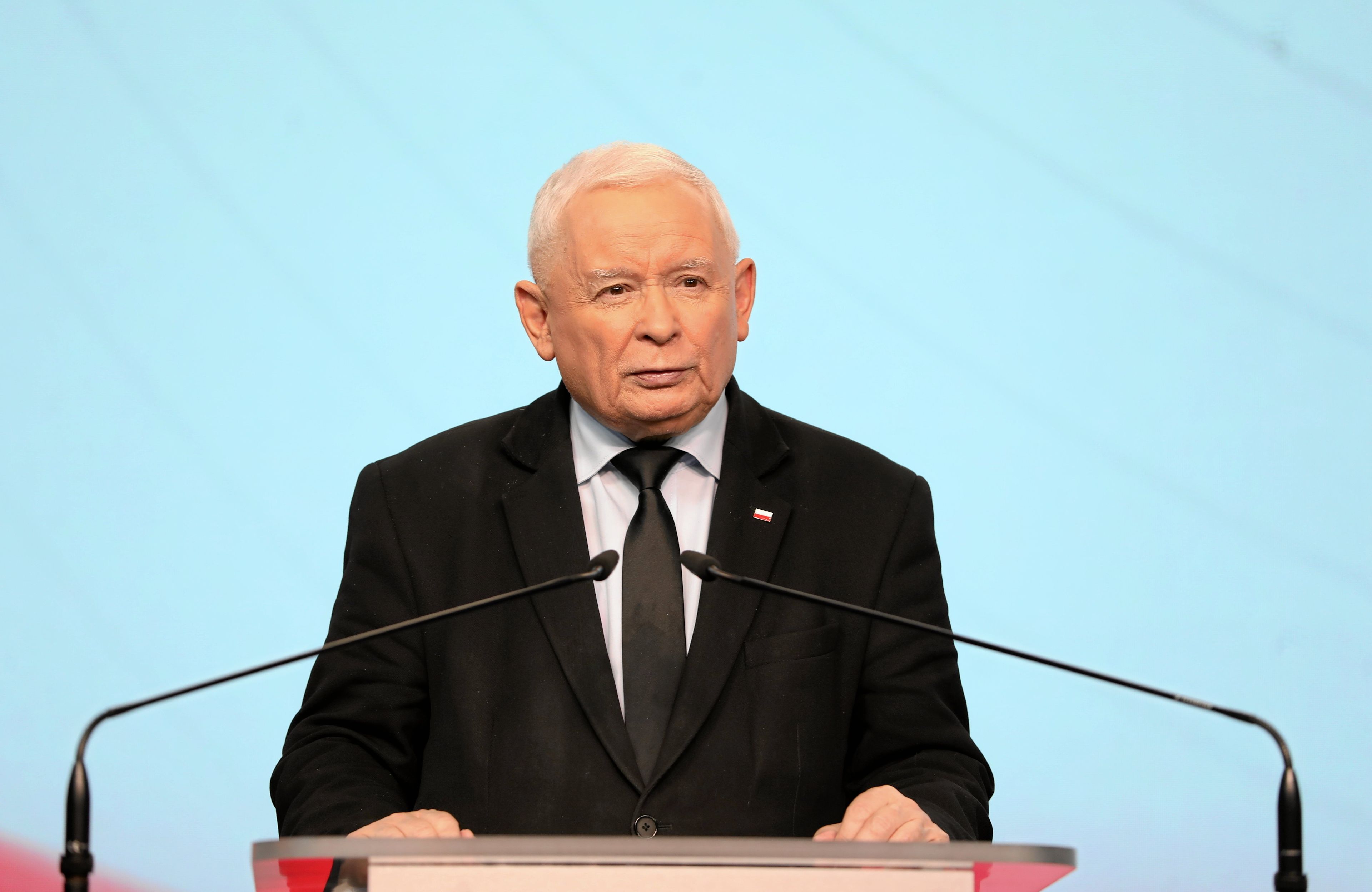 Jarosław Kaczyński w czarnym garniturze i krawacie. Przemawia zza podium na konferencji prasowej.