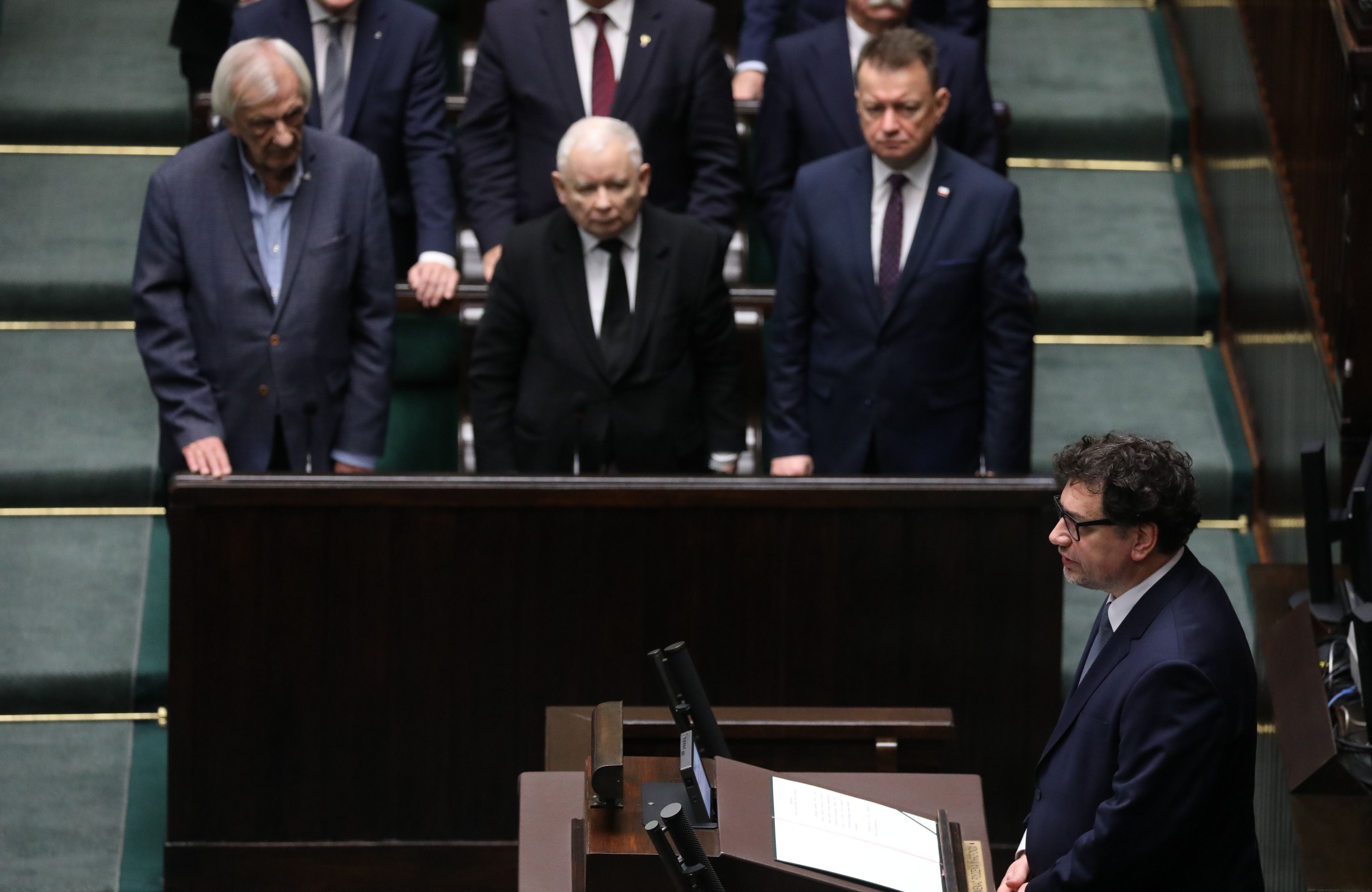 Męzczyzna przy mównicy sejmowej. Posłowie PiS stoją