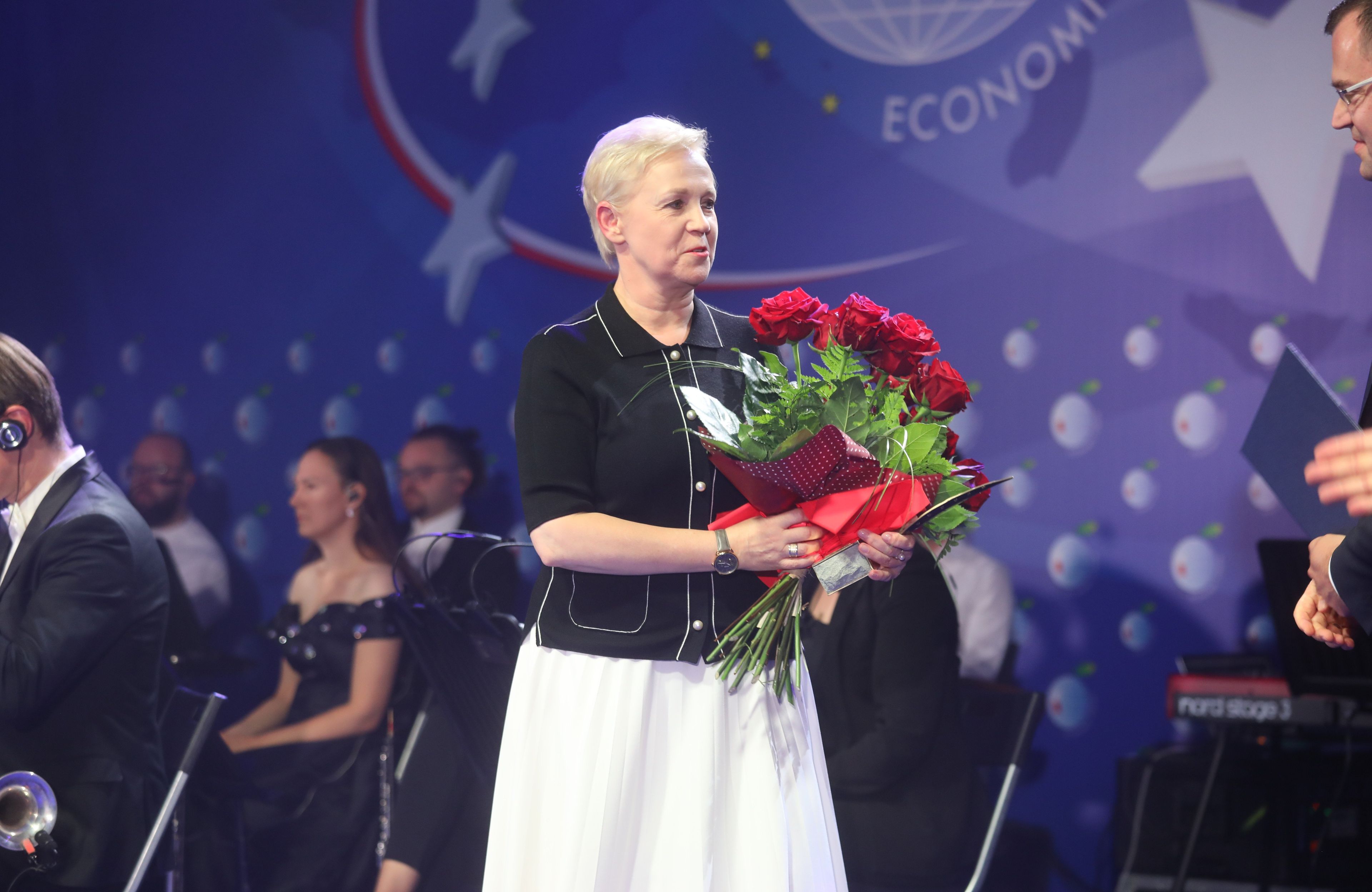 Kobieta, blondynka, w wieczorowym stroju na scenie, w rękach trzyma kwiaty