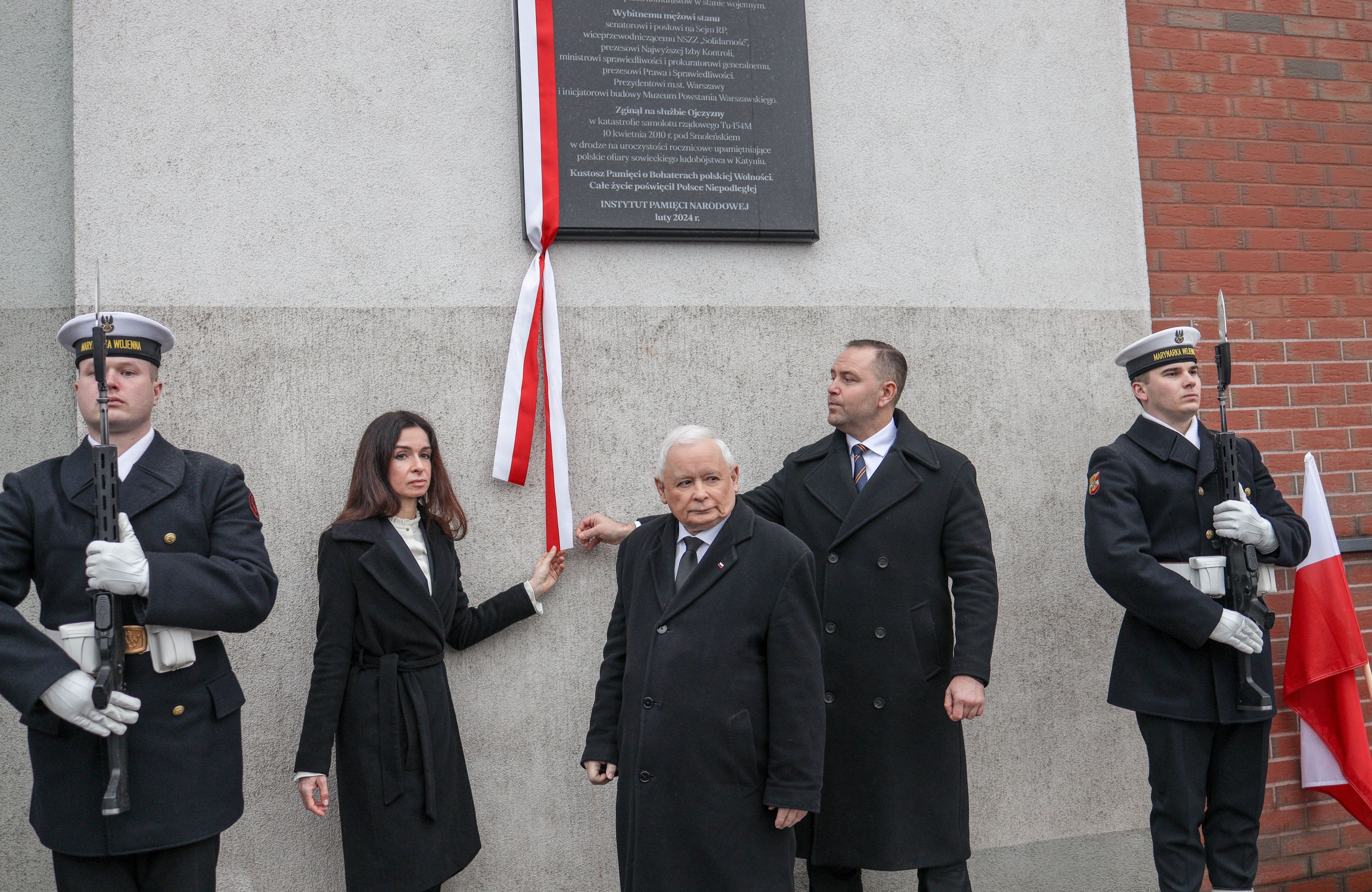 Kilka osób stoi pod wmurowaną w ścianę tablicą upamiętniającą