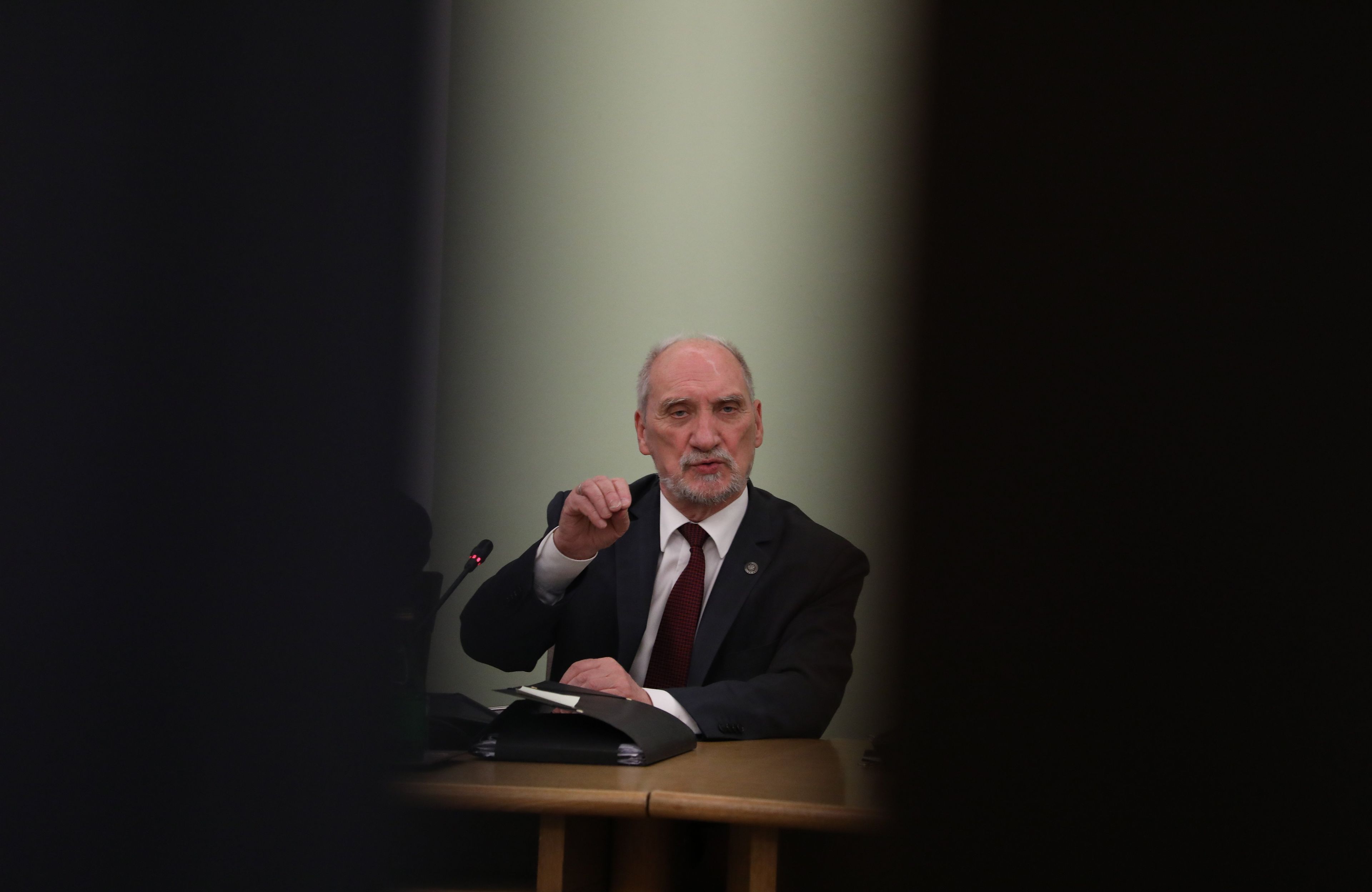 Mężczyzna w garniturze siedzi przy stole prezydialnym, zdjęcie zrobione zza uchylonych drzwi