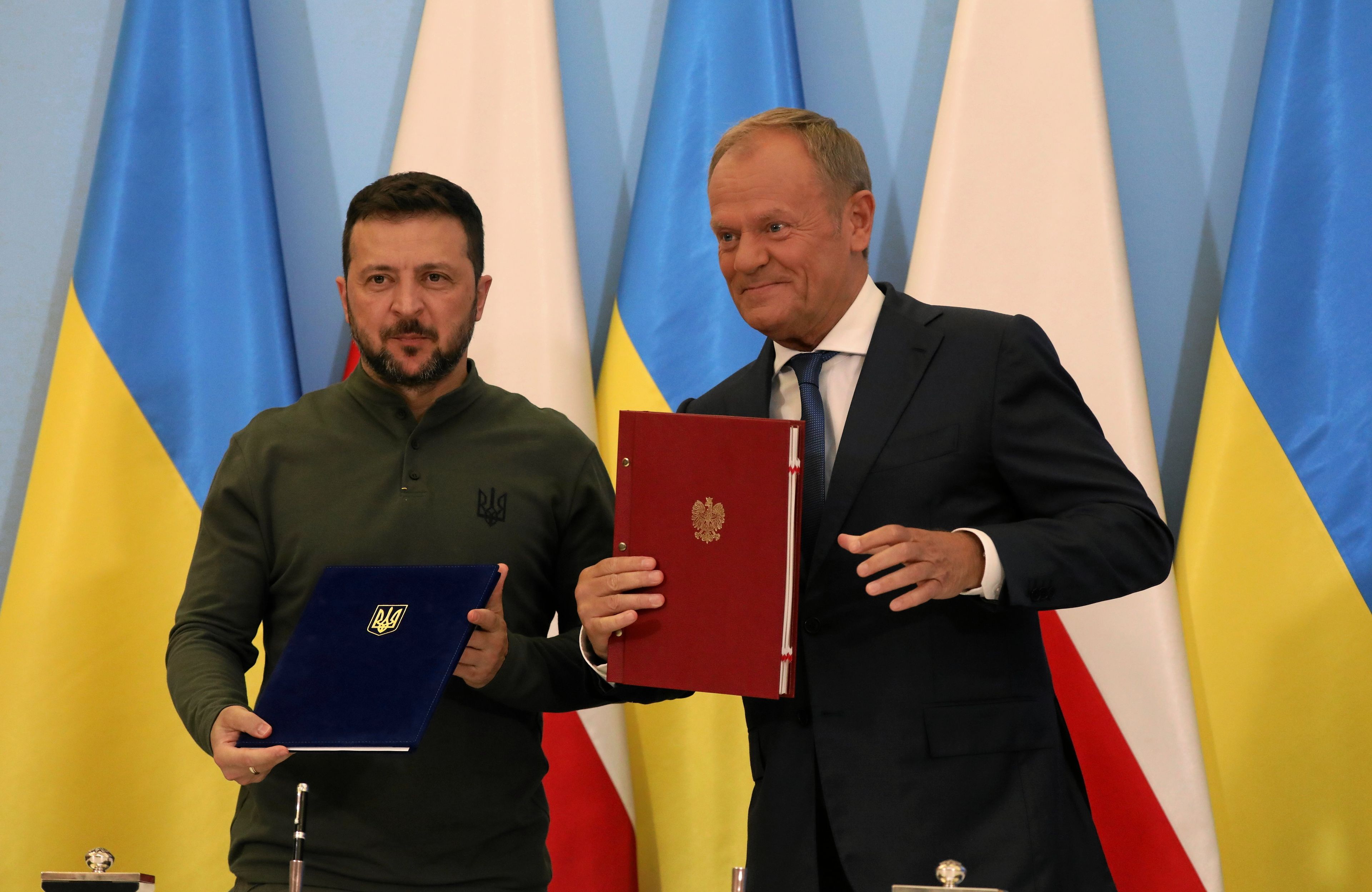 Na zdjęciu jest dwóch mężczyzn. Mężczyzna po lewej ok. 45 lat, jest ubrany w bluzę o kolorze khaki, ma czarne włosy i krótką brodę, w ręckach trryma granatową teczkę z godłem Ukrainy. Po prawej mężczyna wygląda na ok 60 lat. Jest w czarnym garniturze i białej koszuli, ma jasne włosy. Trzyma czerwoną teczkę z godłem Polski. Z tyłu są flagi niebiesko-żółte oraz biało-czerwone