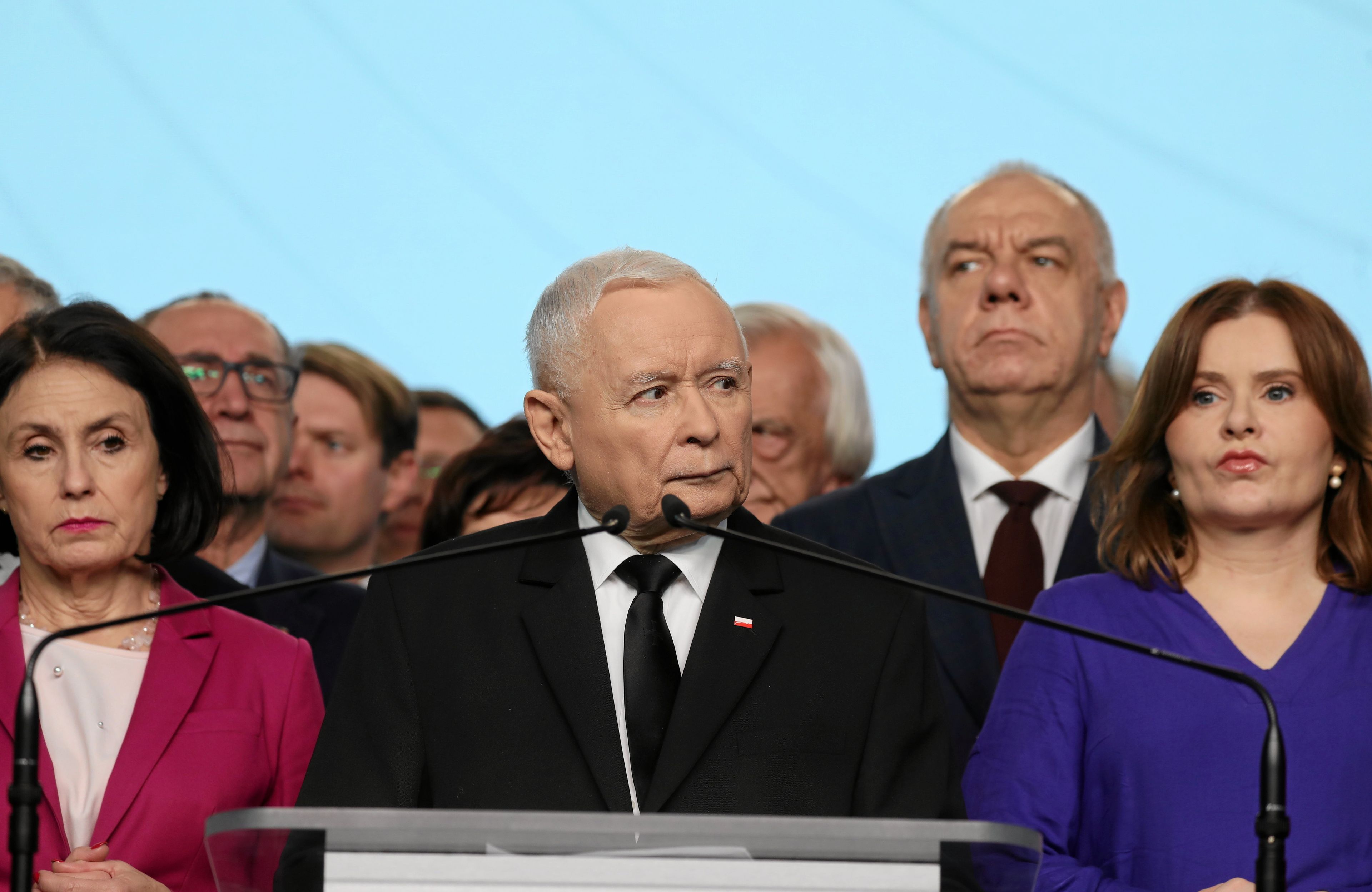 Na pierwszym planie starszy, siwy mężczyzna. Po jego prawej i lewej stronie kobiety, z tyłu łysawy mężczyzna wyciąga głowę