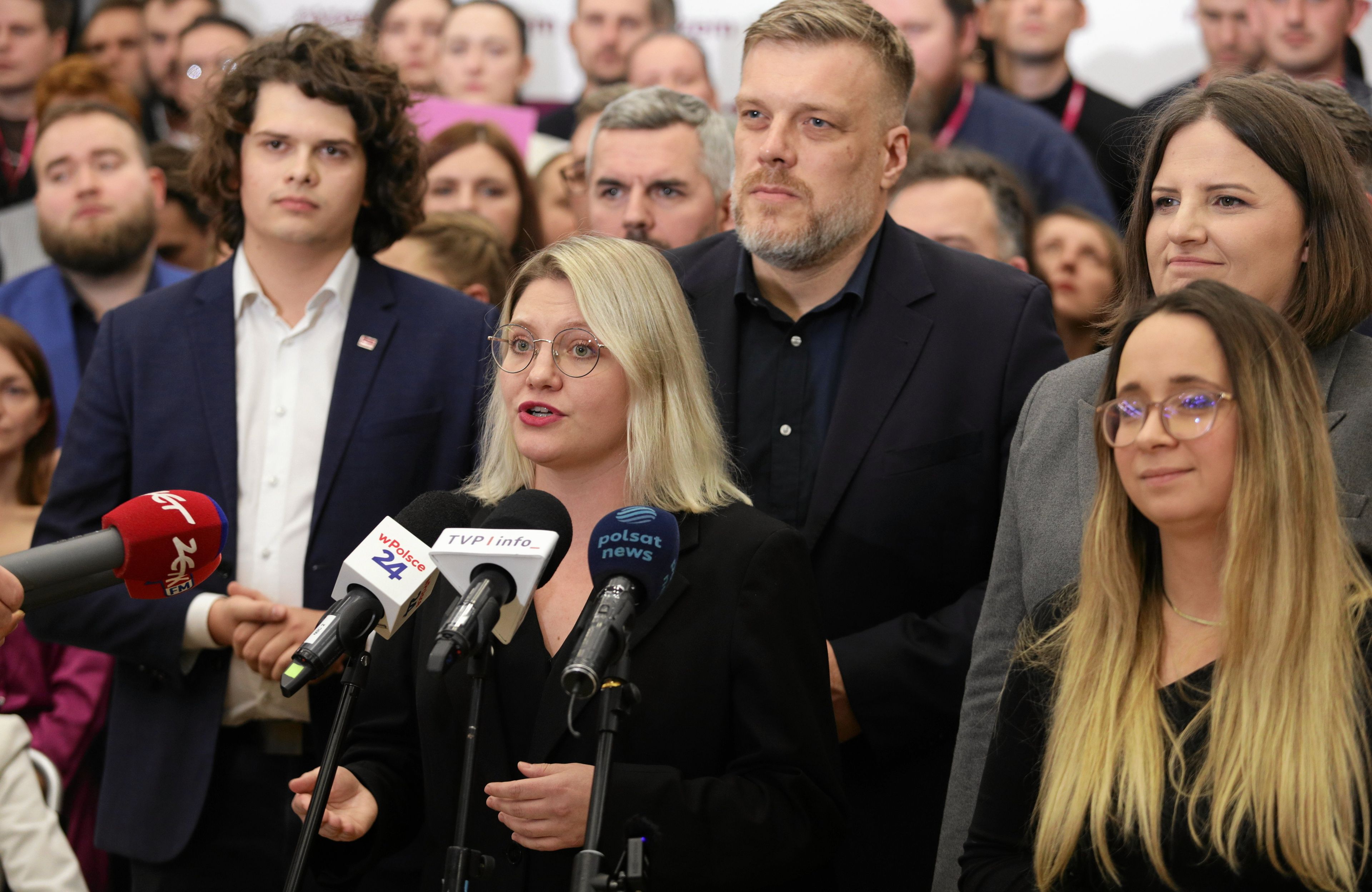 wiele młodych osób, na pierwszym planie blondynka w okularach przed mikrofonem, za nią potężny mężczyzna w ciemnym stroju