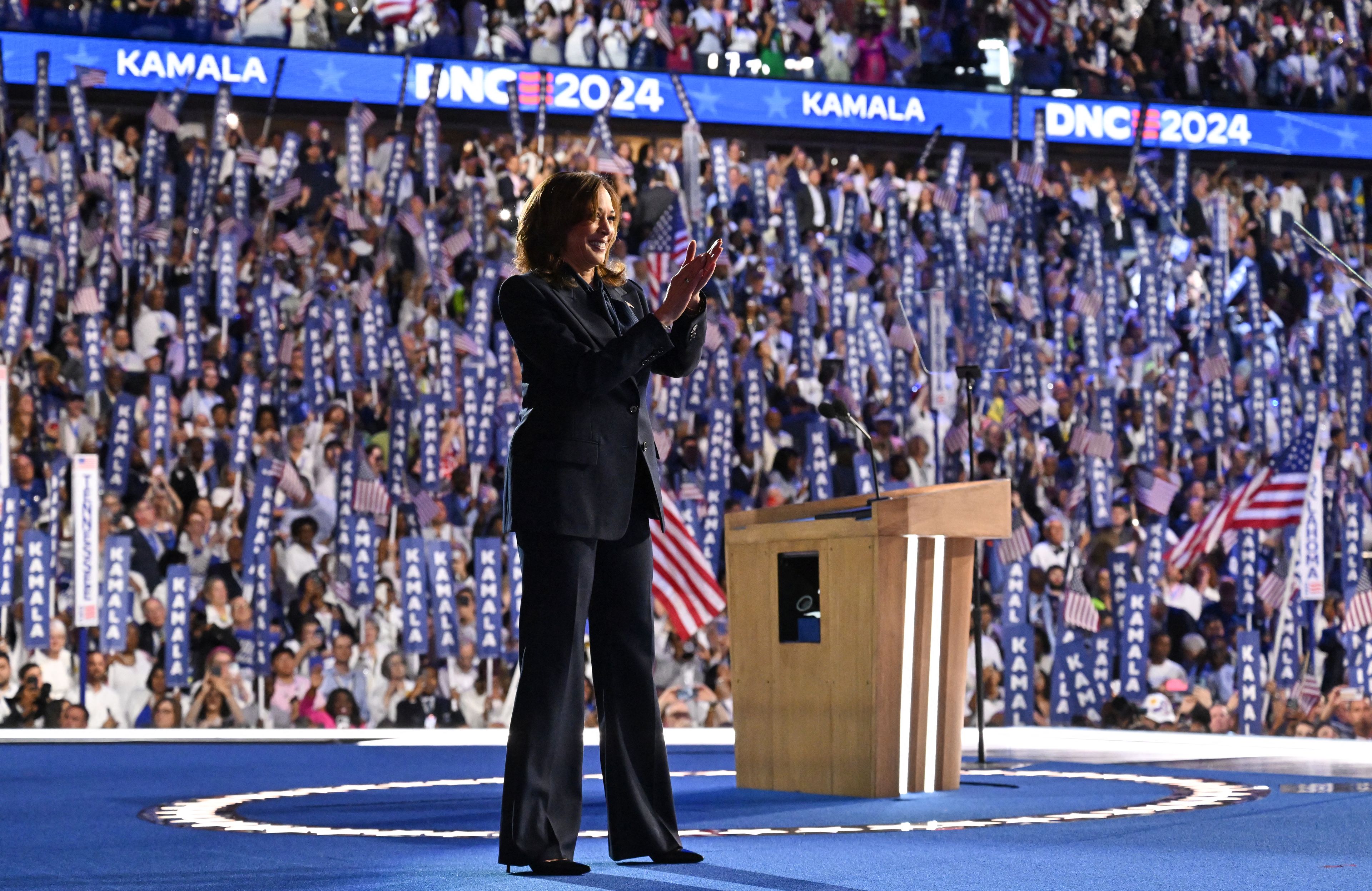 Kamala Harris stoi na dużej scenie i pozdrawia tłumy zwolenników zebranych w hali widowiskowej w Chicago