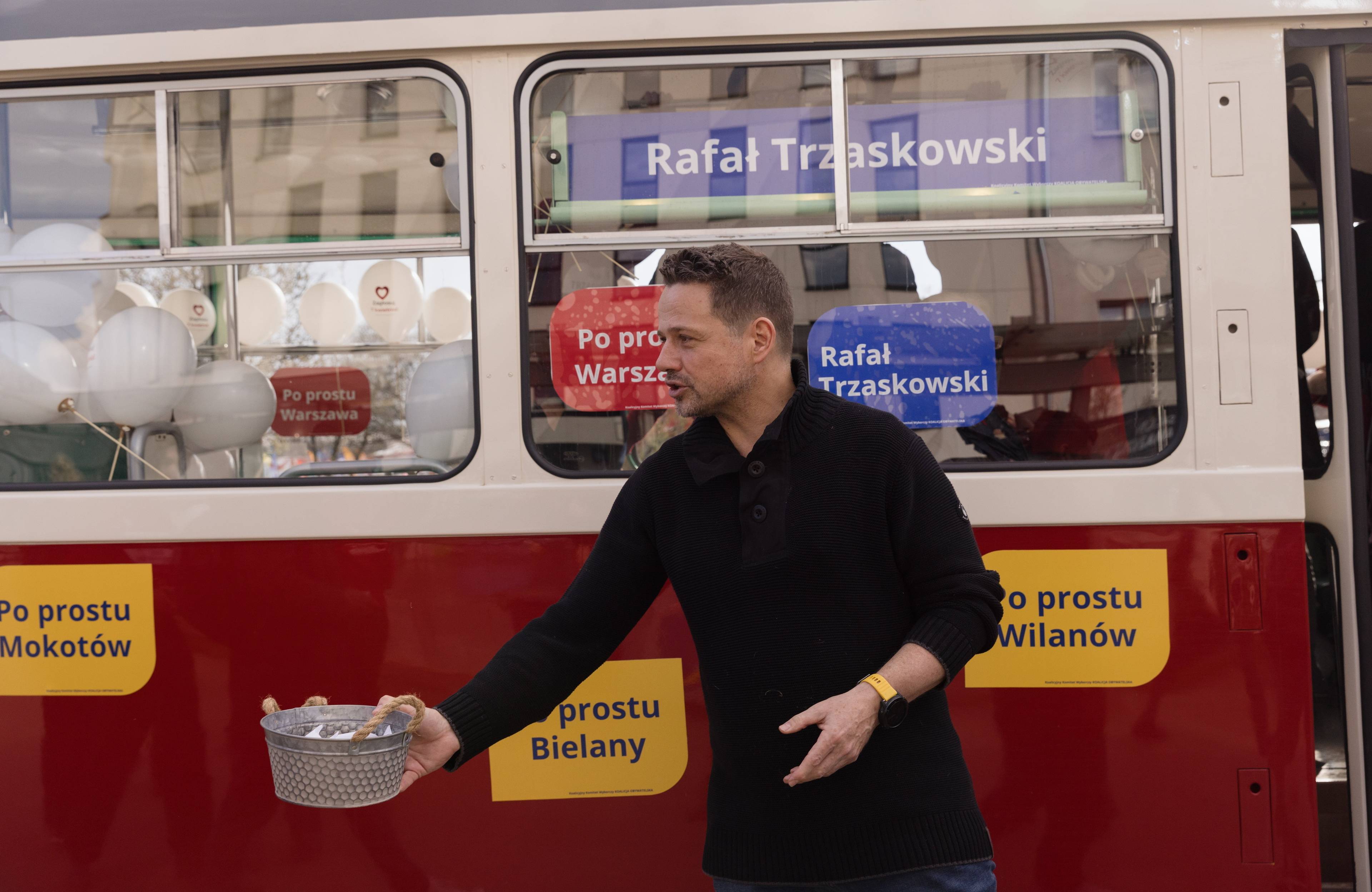 Rafał Trzaskowski rozdaje cukierki przed tramwajem oklejonym hasłami jego kampanii wyborczej