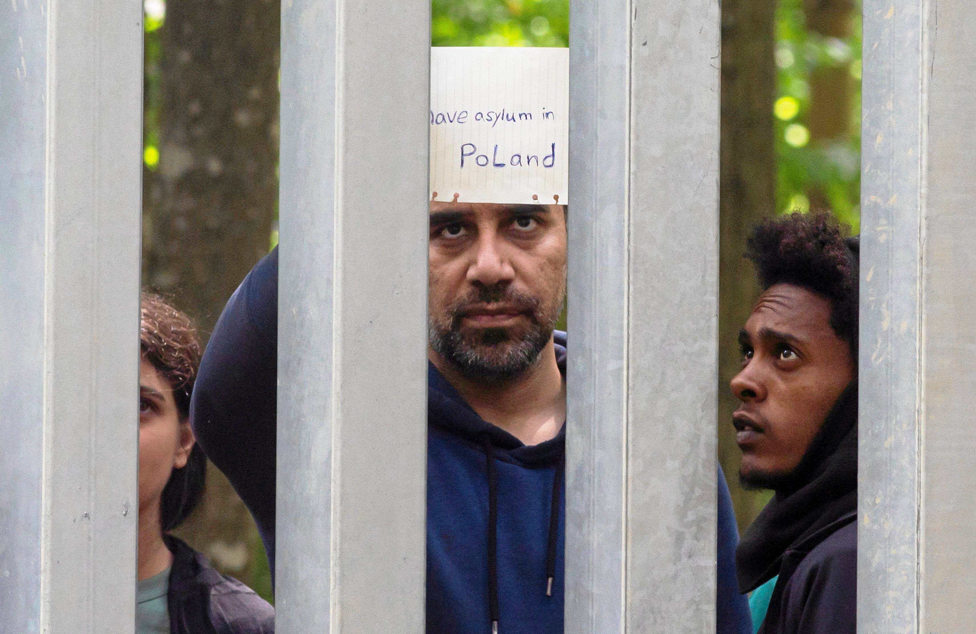Płot graniczny na polsko-białoruskiej granicy. Za metalowa zaporą gromadzą się uchodźcy. Mają kartki z napisem: prosimy o azyl