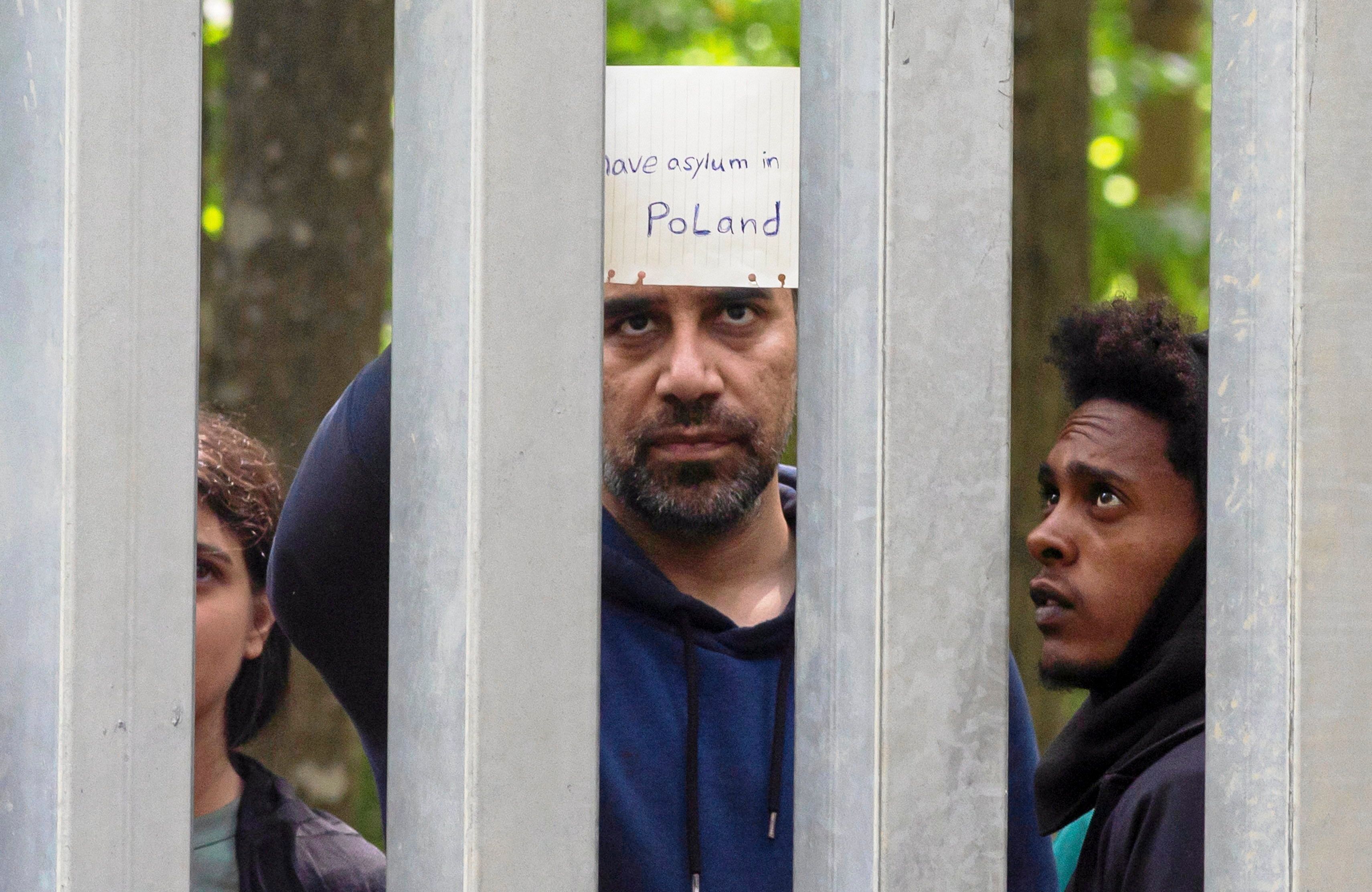 Płot graniczny na polsko-białoruskiej granicy. Za metalowa zaporą gromadzą się uchodźcy. Mają kartki z napisem: prosimy o azyl