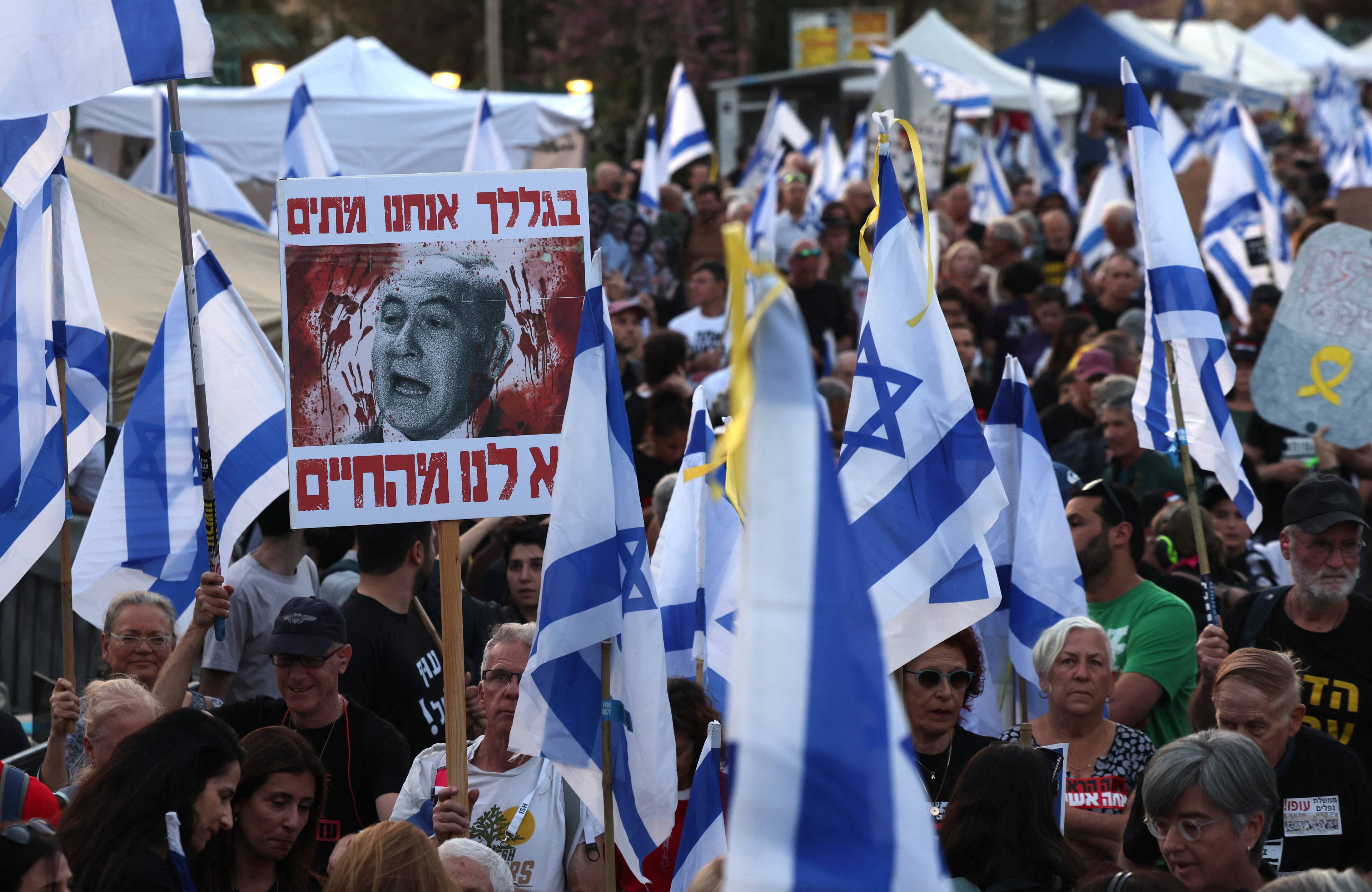 Protest w Izraelu, nad tłumem izraelskie flagi i portret Beniamina Netanjahu z domalowanymi śladami krwi