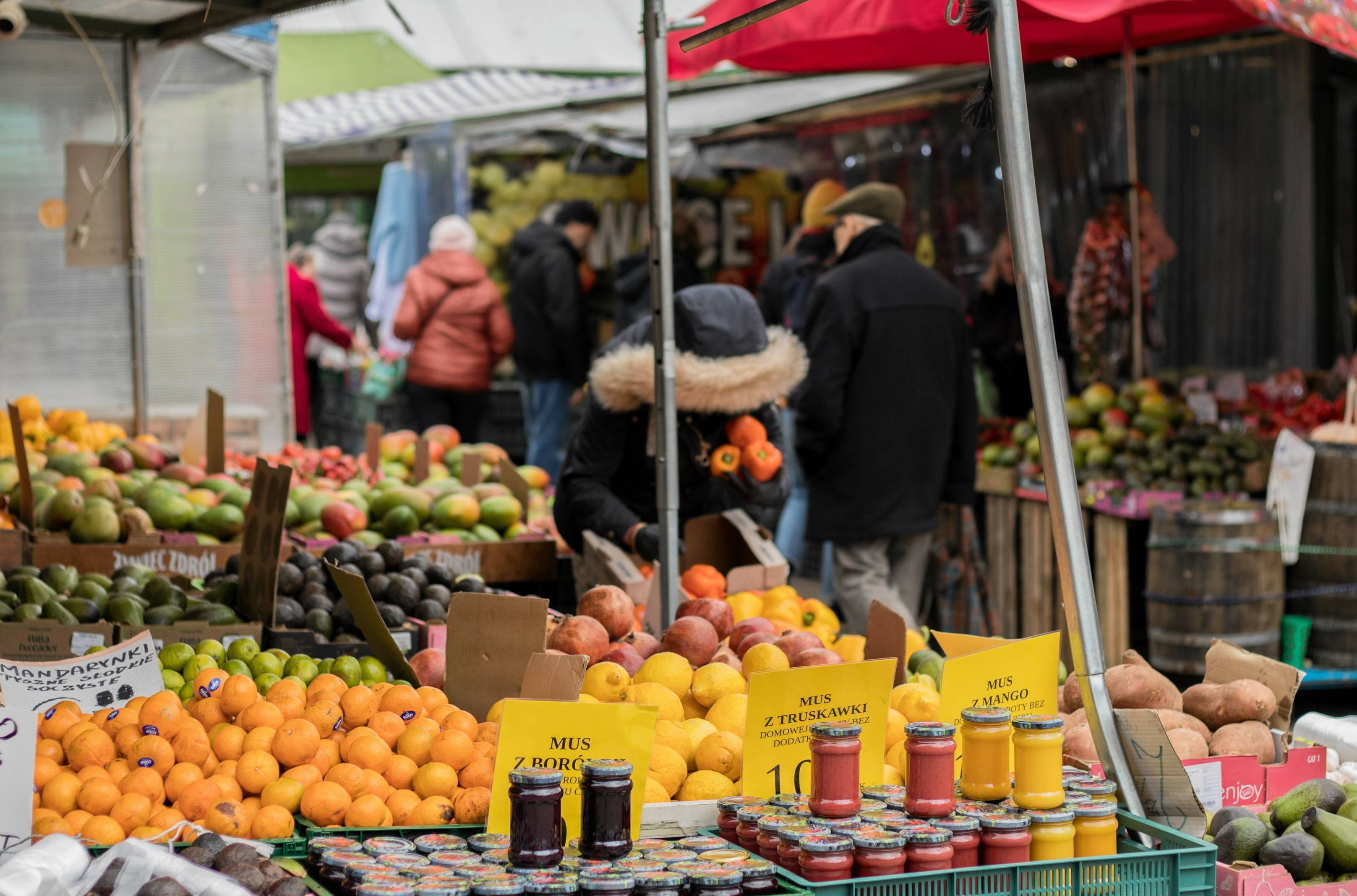 Kobieta pochylona nad owocami i przetworami na bazarze, patrzy na ceny