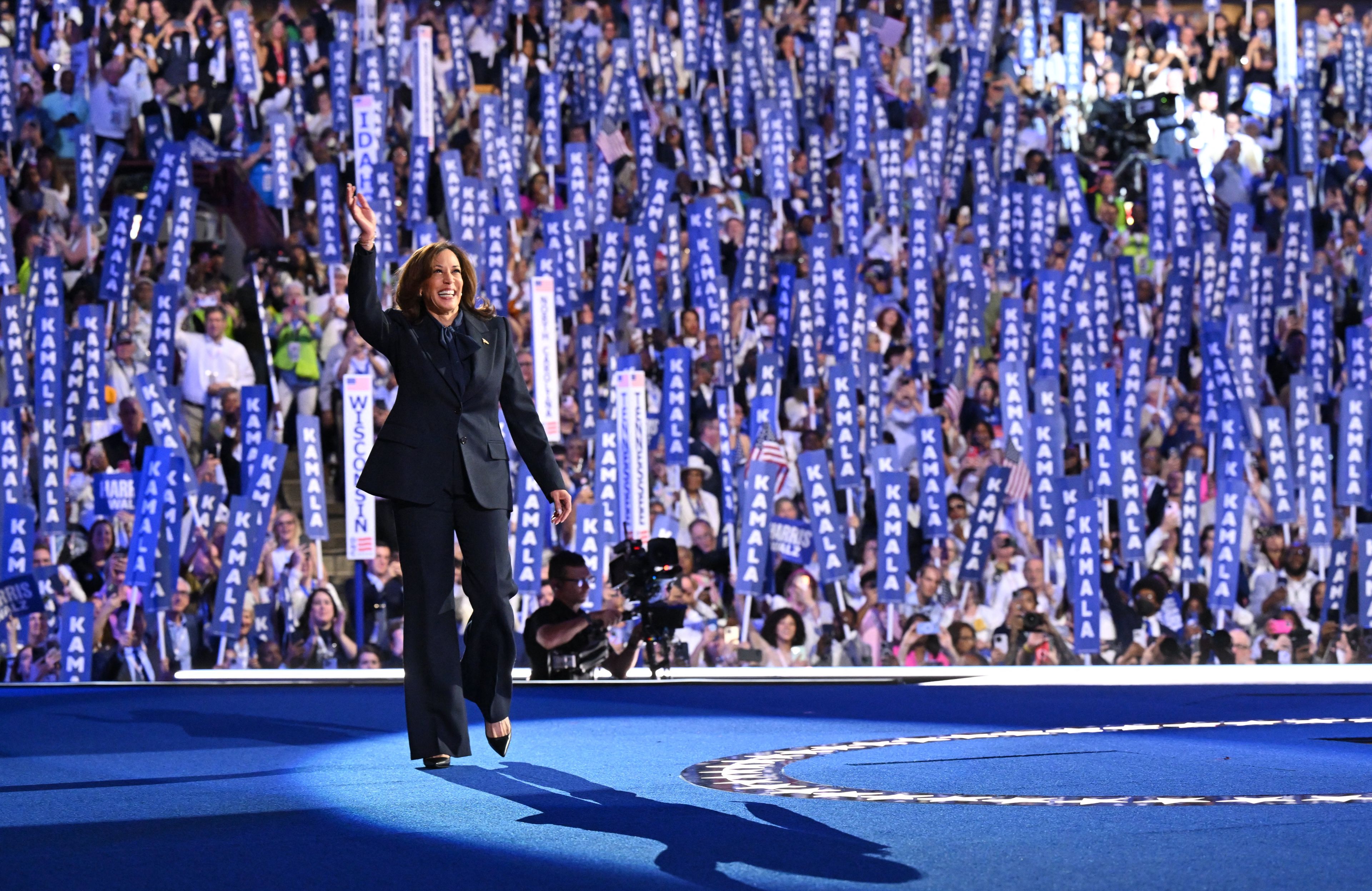 kamala harris w garniturze na tle tłumu, trzymającego napisy "kamala"