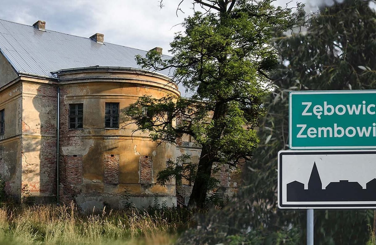 Zielona tablica z podwójną nazwą miejscowości: Zębowice i Zembowitz, w tle niszczejący pałac wśród zieleni.