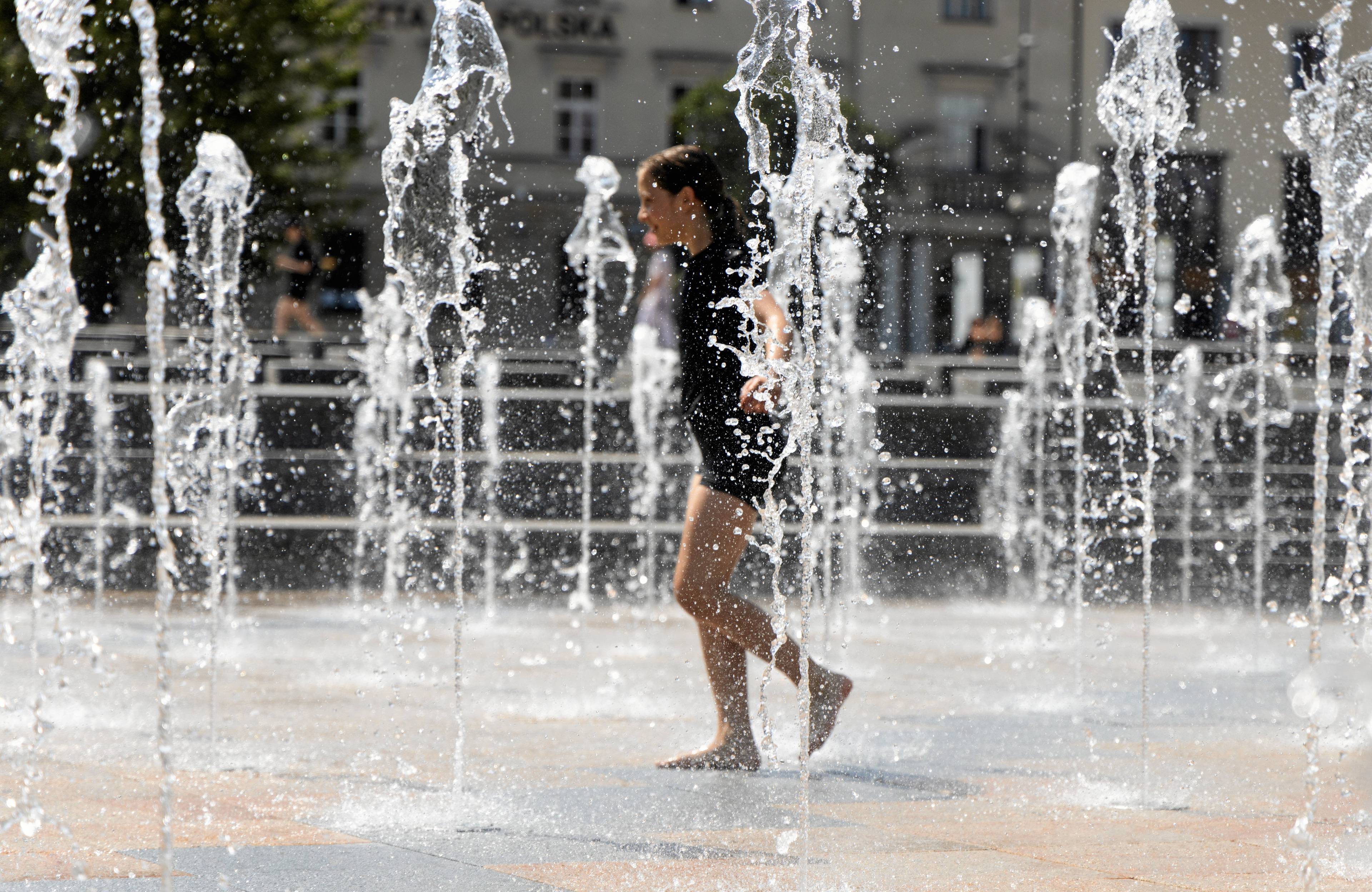 dziewczynka w miejskiej fontannie na środku betonowego placu, słupy wody strzelają do góry