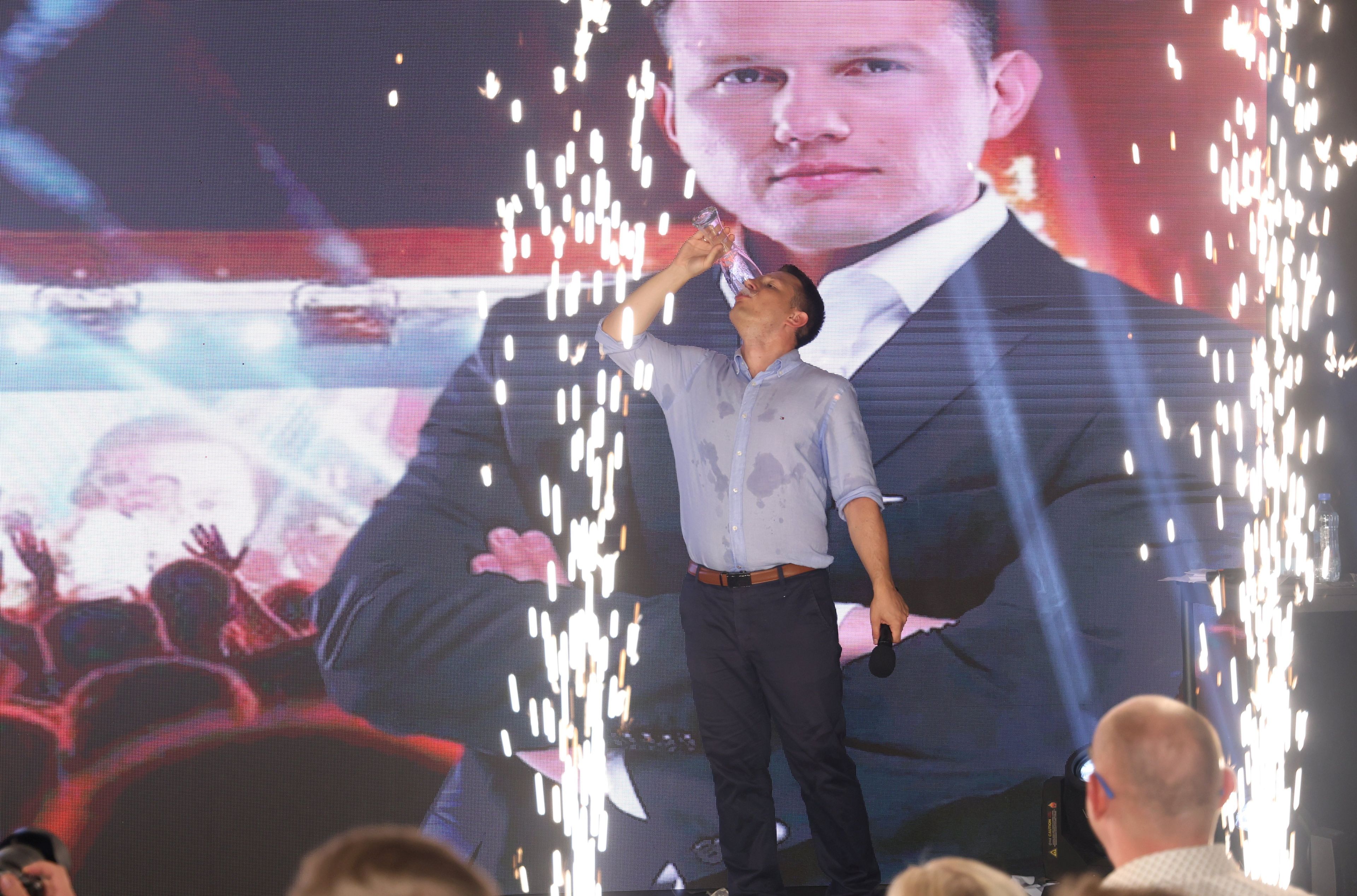 Lider formacji Konfederacja Sławomir Mentzen przechyla szklankę z piwem, na błękitnej koszuli ma plamy, za nim duży banner z jego własną podobizną