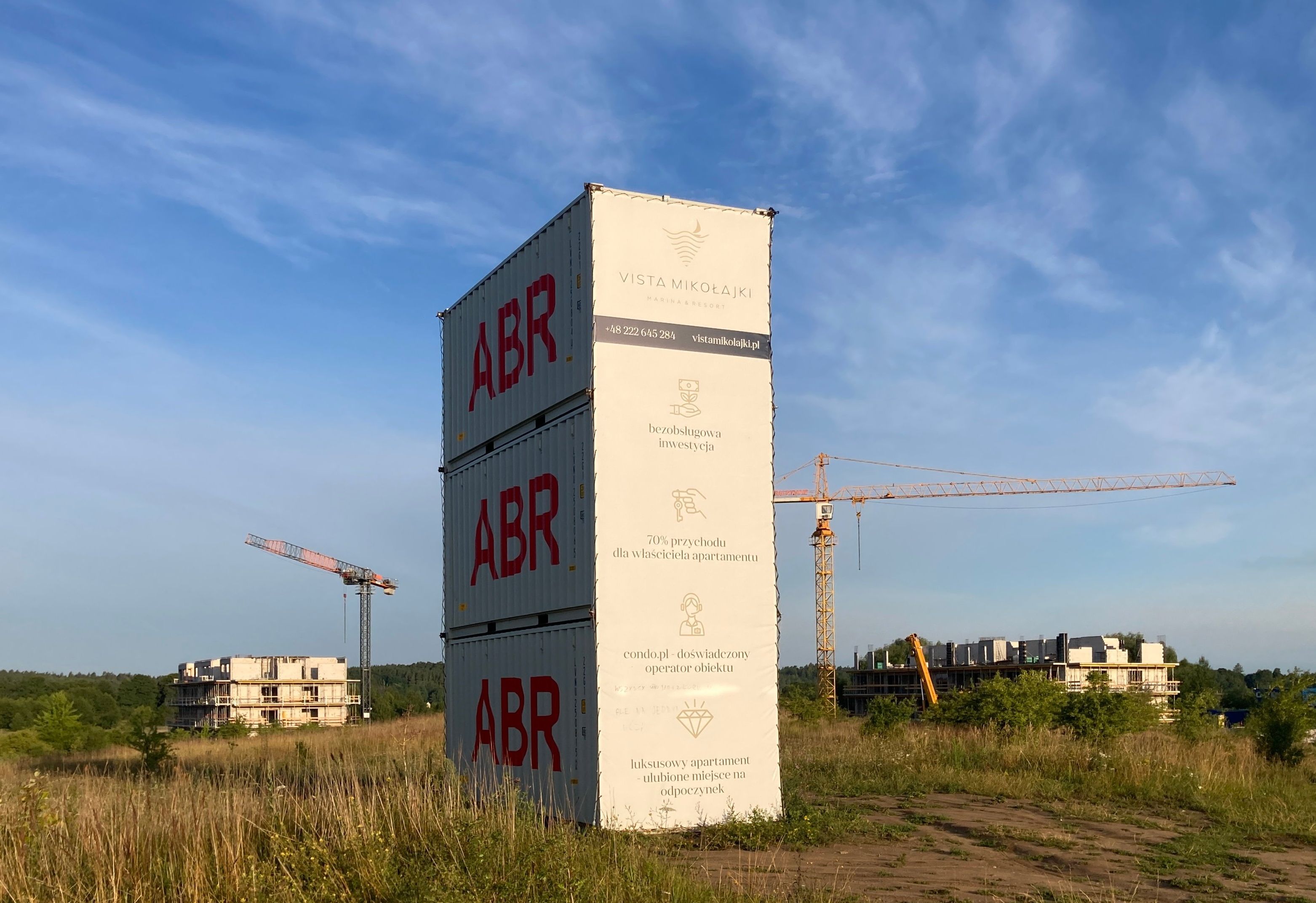 Trzy kontenery budowlane ustawione piętrowo, dźwigi i budowa bloków wśród łąk, w tle las