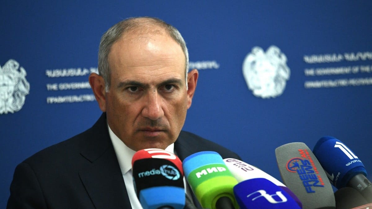 Łysawy mężczyzna w garniturze wsłuchuje się w pytanie dziennikarza, w tle herb Republiki Armenii