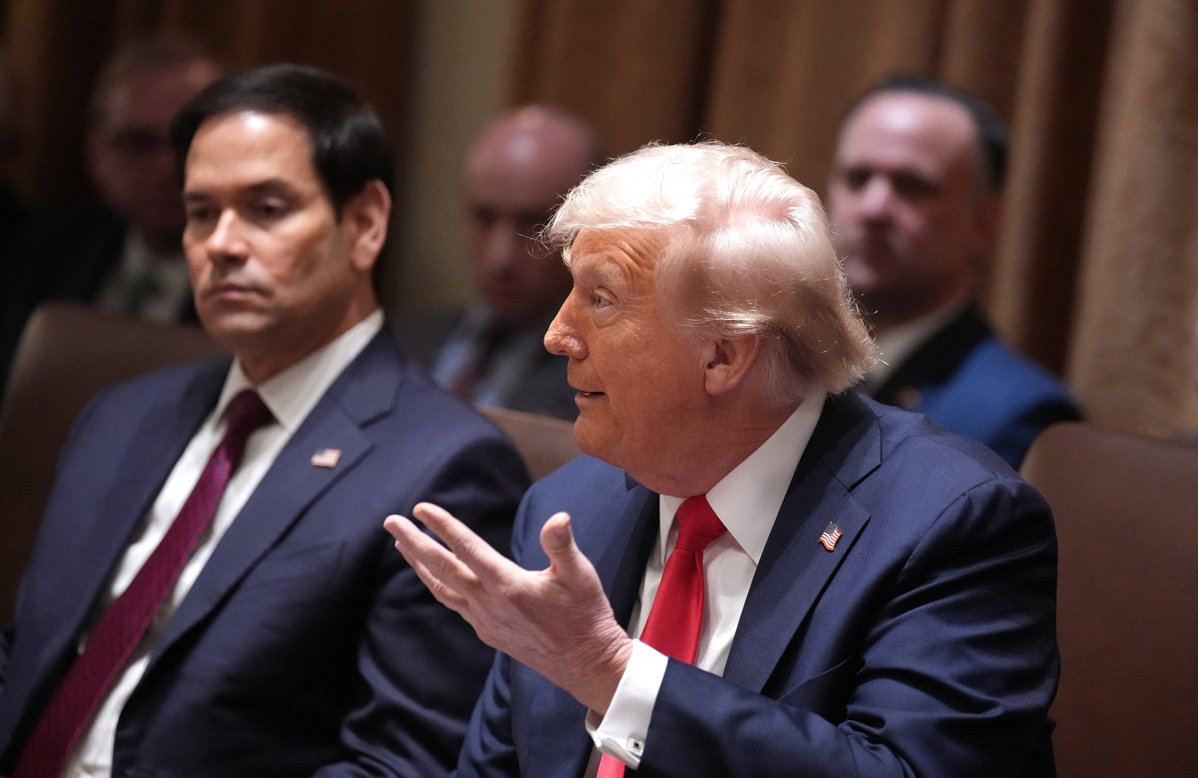 Mężczyzna z jasnymi rzadkimi włosami, Donald Trump, przemawia siedząc przy dużym owalnym stole
