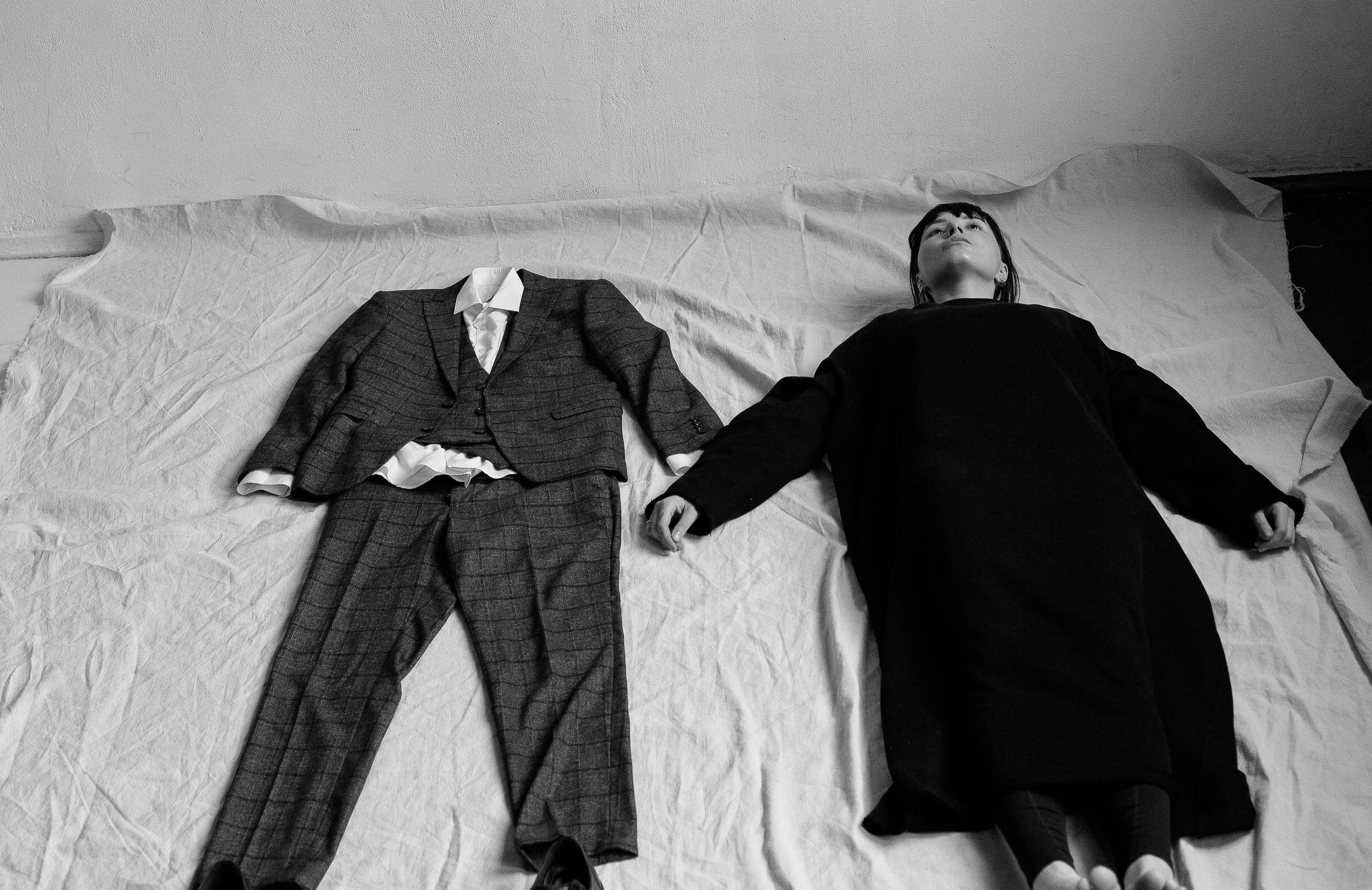 Kobieta leży na łóżku z rozłozonymi ramionami obok garnituru mężczyzny. Zdjęcie czarno-białe
