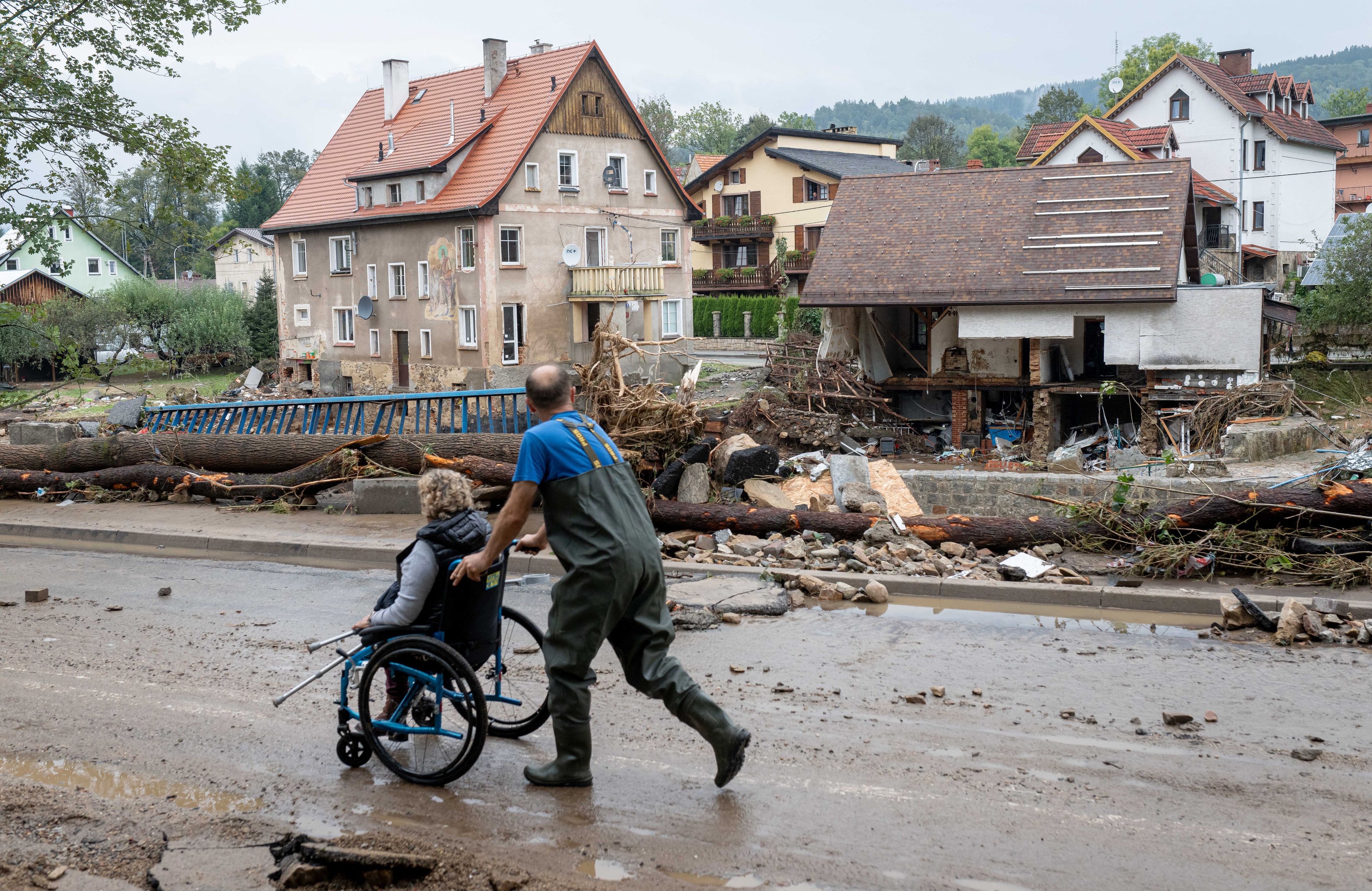 Mężczyzna pcha kobietę na wózku dla osób niepełnosprawnych. Wokół miasto zniszczone przez powódź