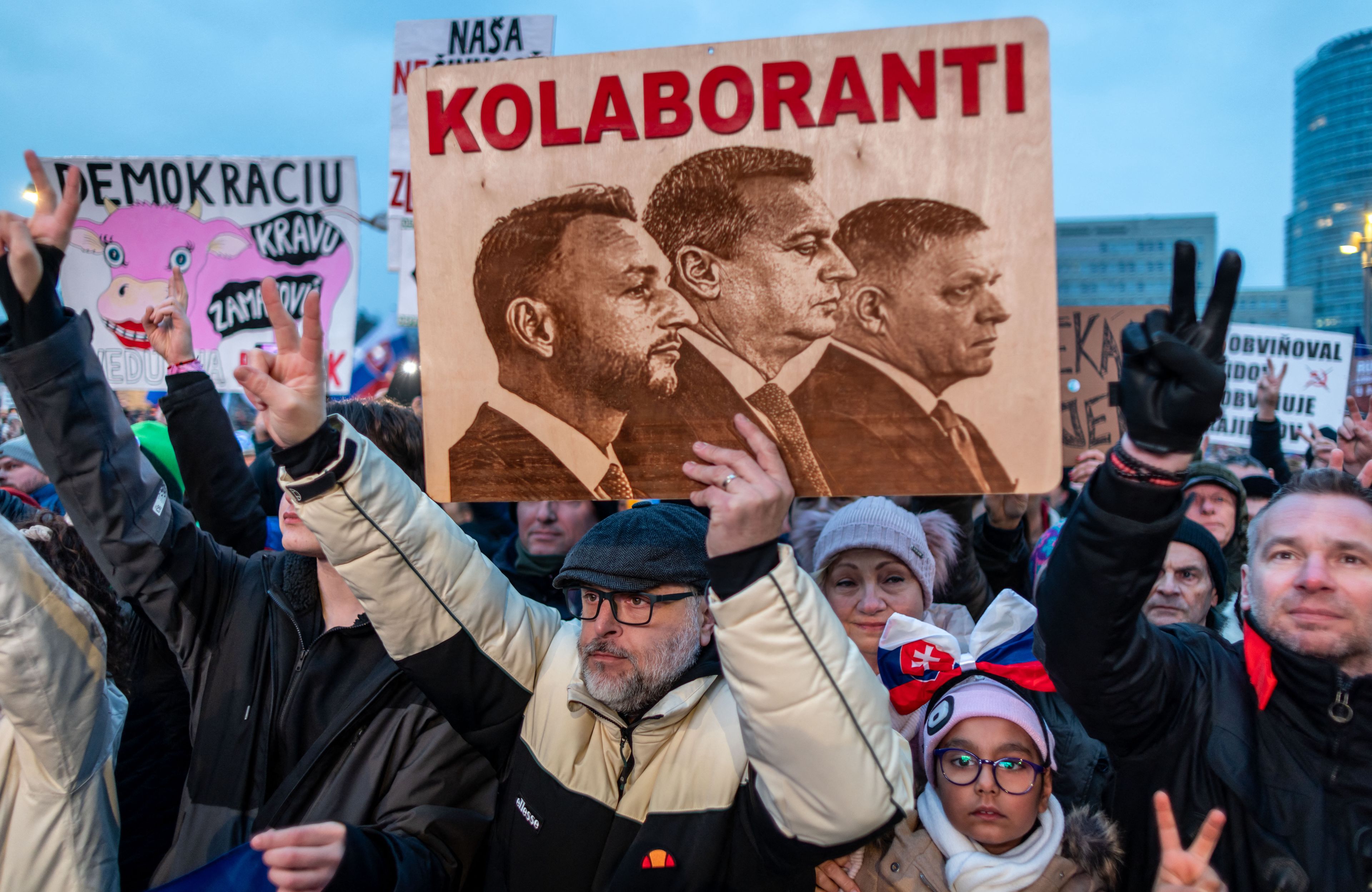 Brodaty mężczyzna stoi w tłumie ludzi. Jedną ręką pokazuje znak zwycięstwa, w drugiej trzymaz transparent przedstawiający trzech mężczyzn w garniturach i napisem "Kolaboranti"