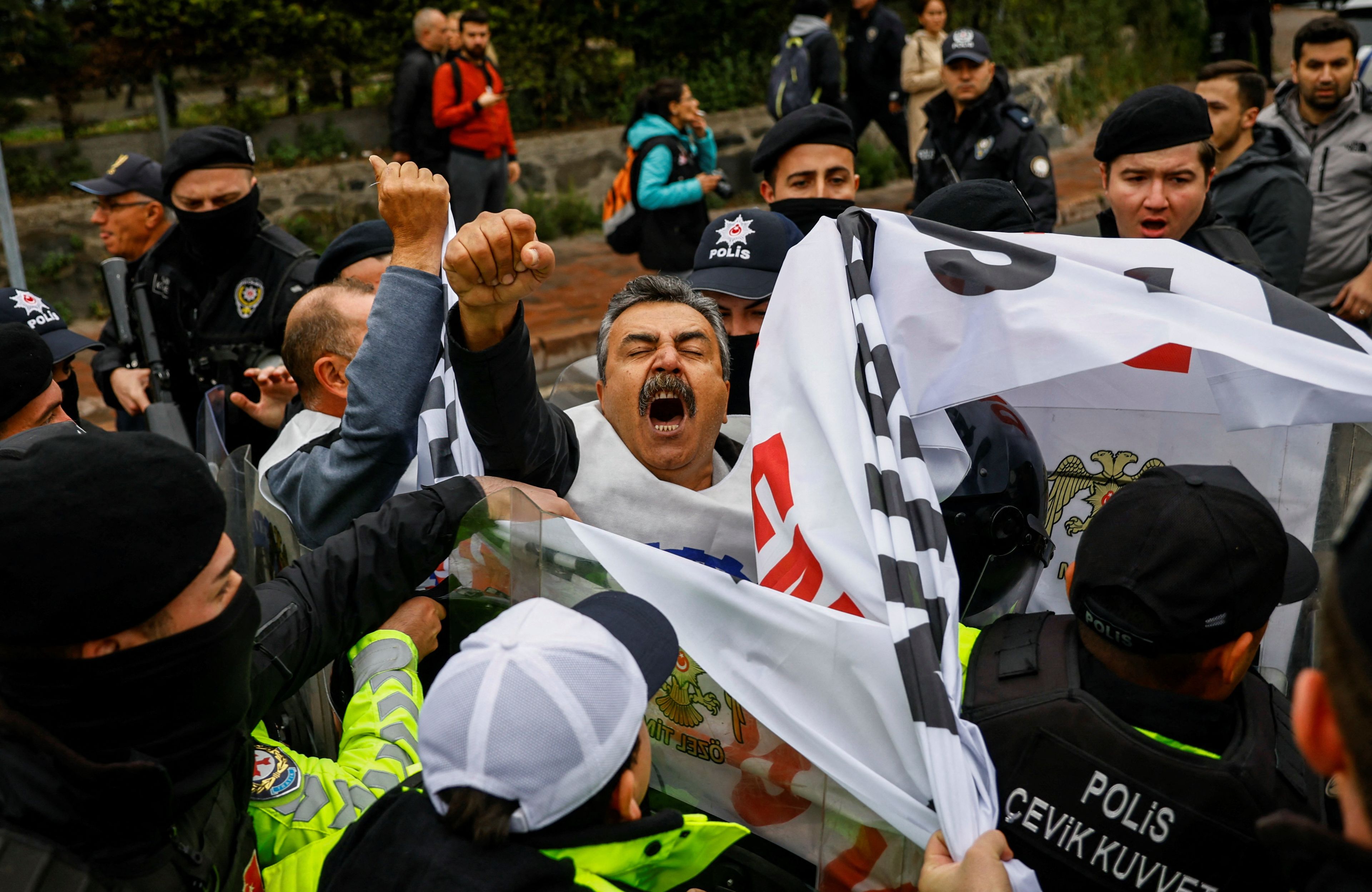 Mężczyzna z czarno-siwymi włosami i wąsami gęsto otoczony przez tłum protestujących i policję wznosi w powietrze pięść w geście protestu, policja usiłuje mu wydrzeć materiałowy transparent z hasłem, którego jednak nie da się odczytać.