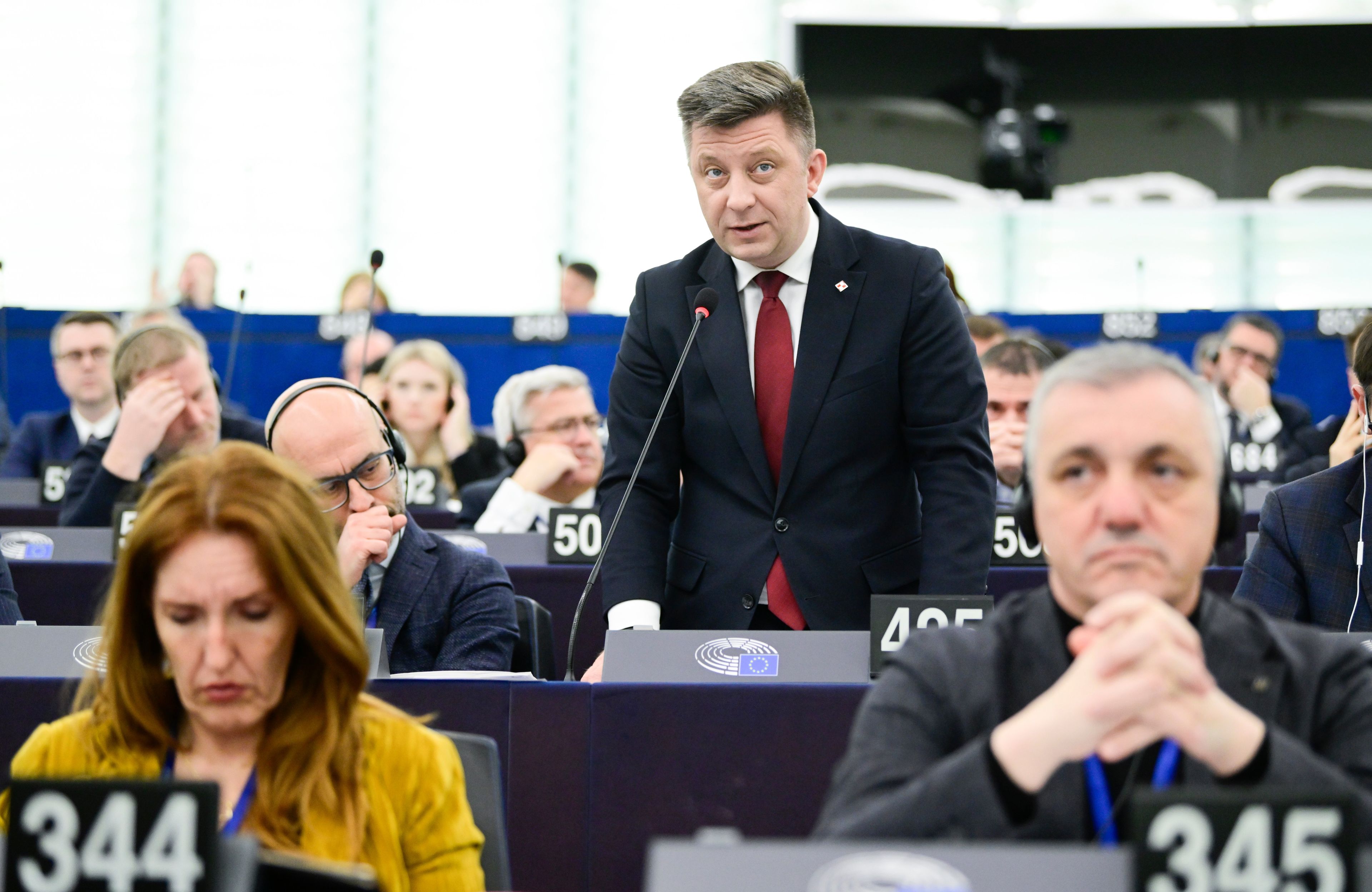 Mężczyzna w ciemnogranatowym garniturze z czerwonym krawatem, z krótkimi ciemnoblond włosami stoi w ławie przy której w rzędzie siedzi więcej osób i mówi do włączonego mikrofonu. Na pierwszym planie, lekko nieostre, dwie kolejne osoby, także siedzące w ławie, po lewej kobieta z długimi brązowymi włosami, po prawej szpakowaty mężczyzna, który opiera podbródek na splecionych dłoniach.