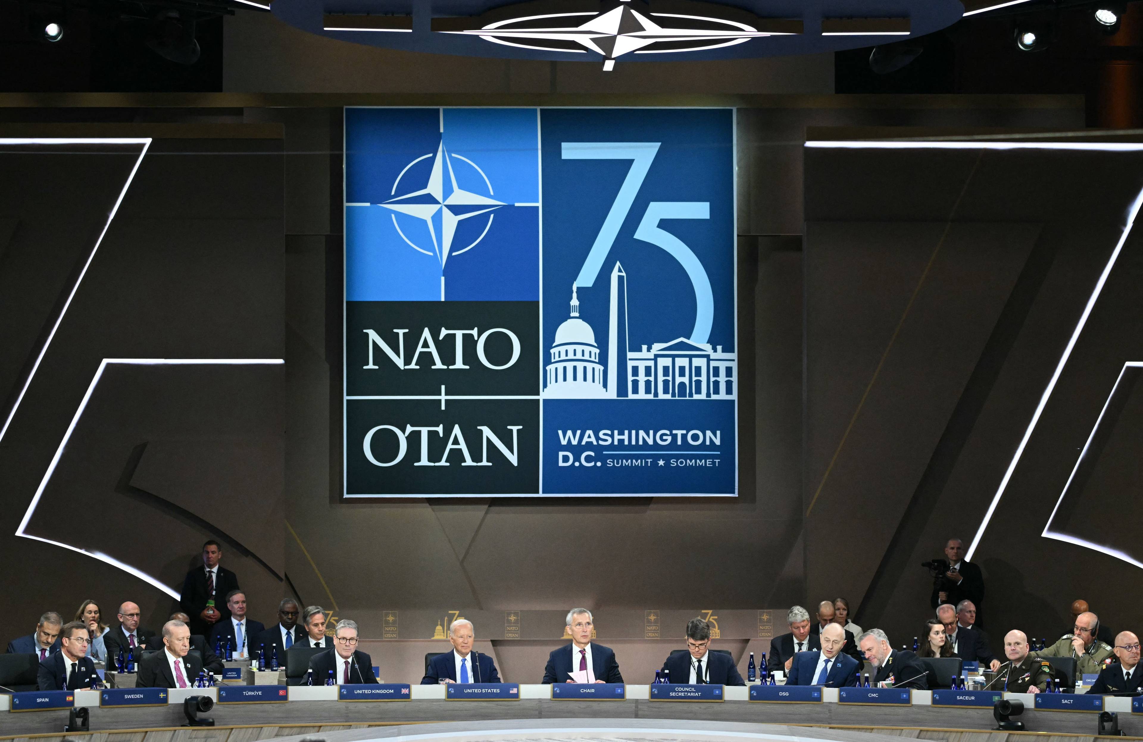 mężczyźni siedzący za stołem pod wielkim logo 75-lecia NATO