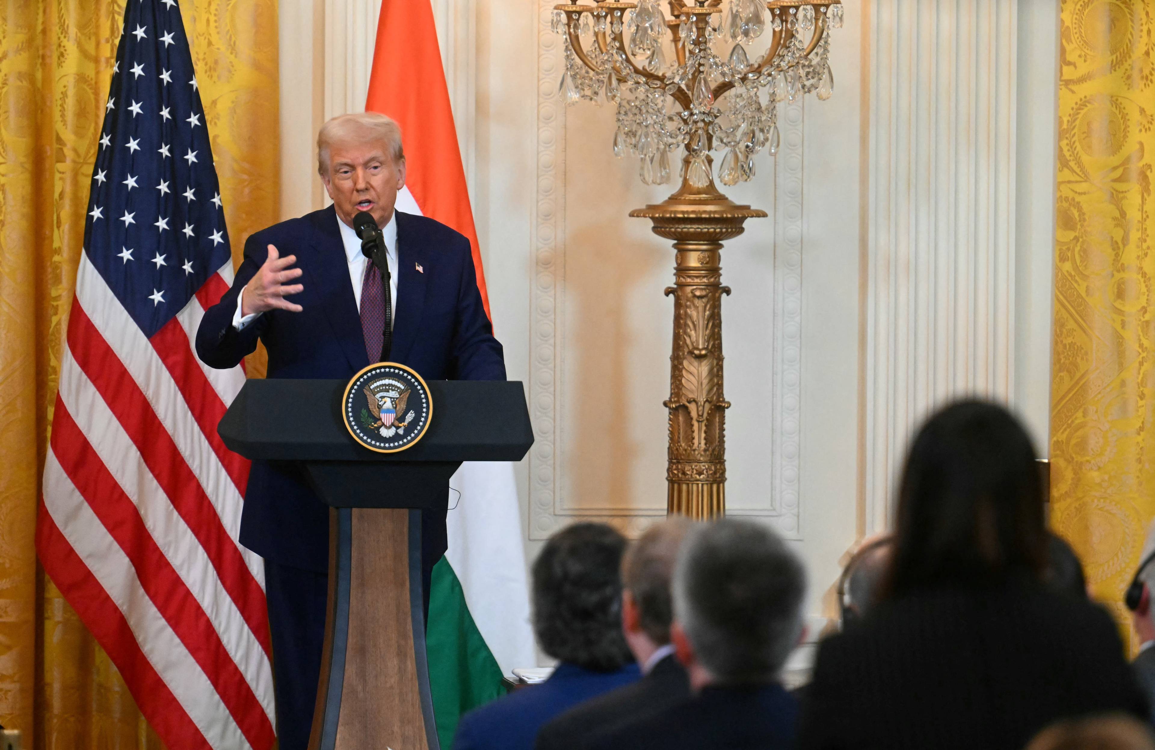 Starszy mężczyzna w garniturze (prezydent USA Donald Trump) przemawia przy pulpicie na tle amerykańskiej flagi