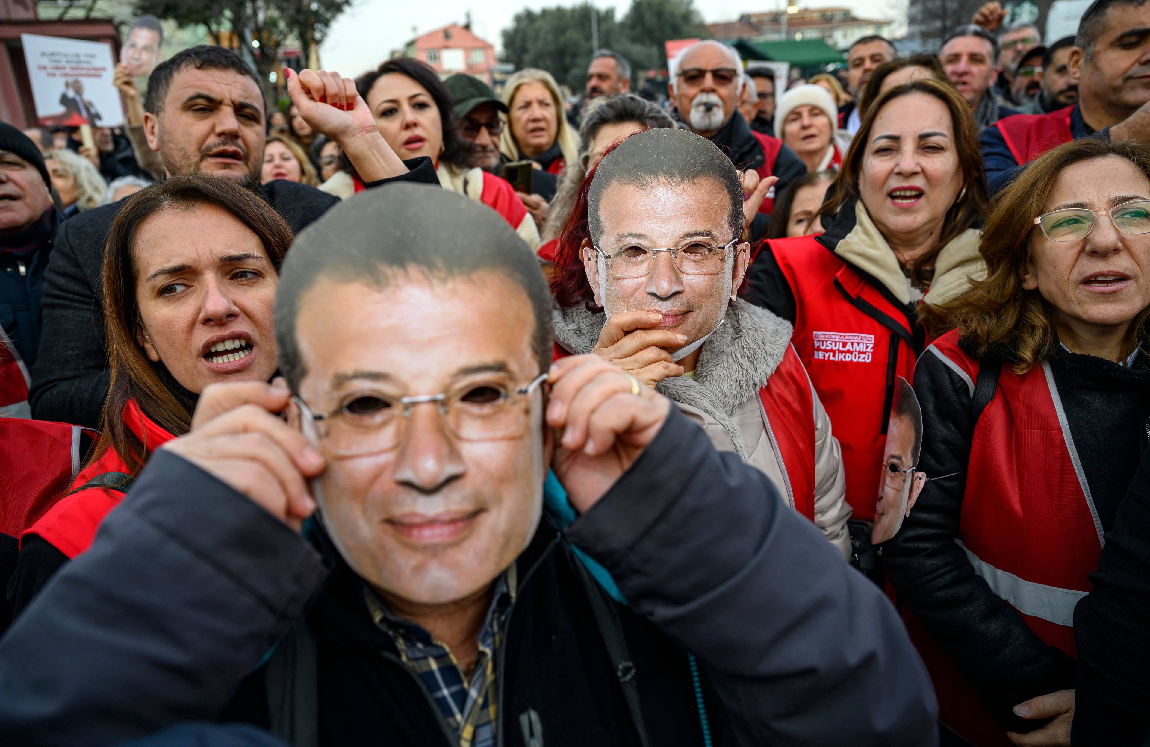 Tłum ludzi, osoba na pierwszym planie trzyma maskę mężczyzny w okularach.