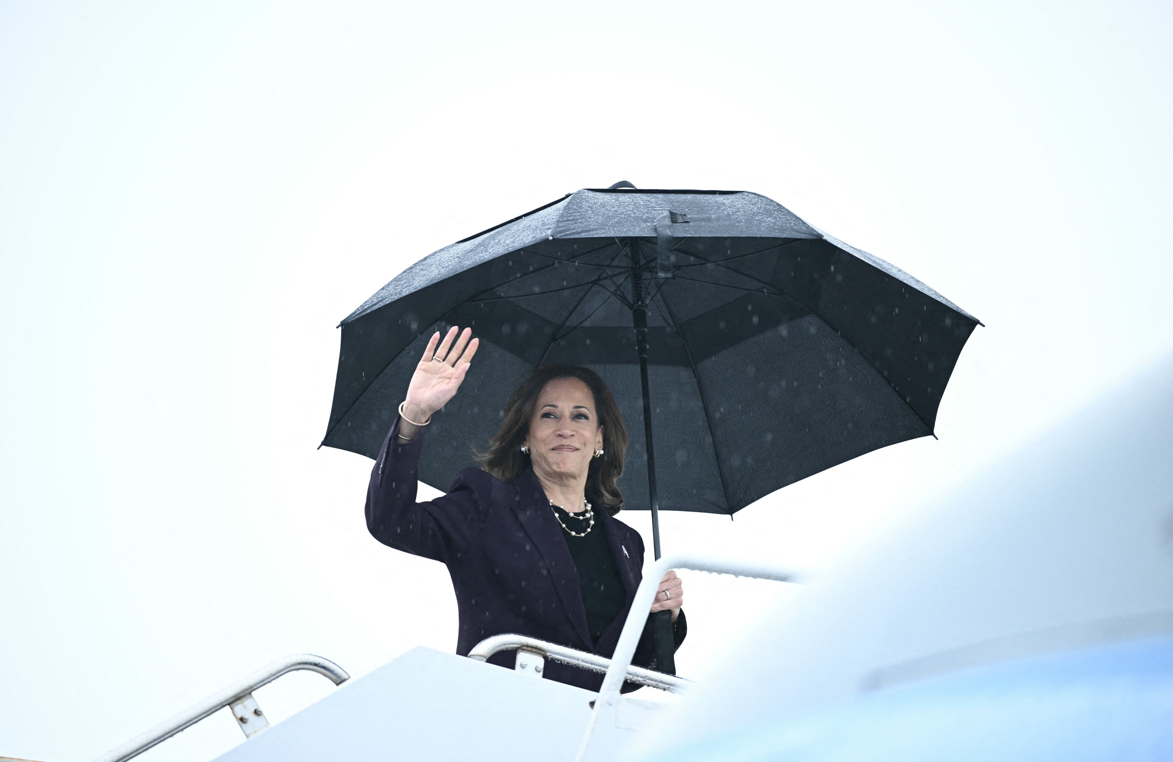 Kamala Harris wchodzi po schodkach do samolotu. W ręku trzyma rozłożony nad głową duży czarny parasol
